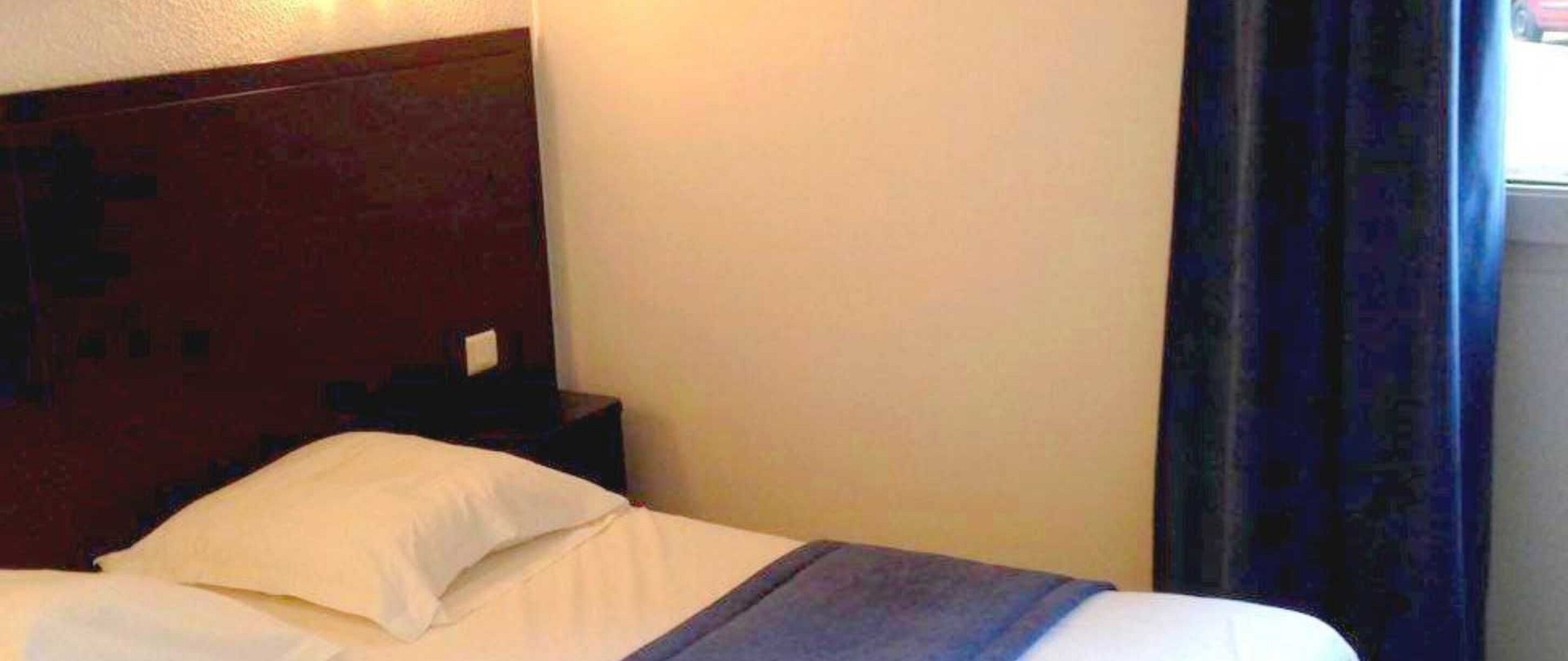 A hotel room with a bed, a wooden headboard, a nightstand, and a window with curtains.