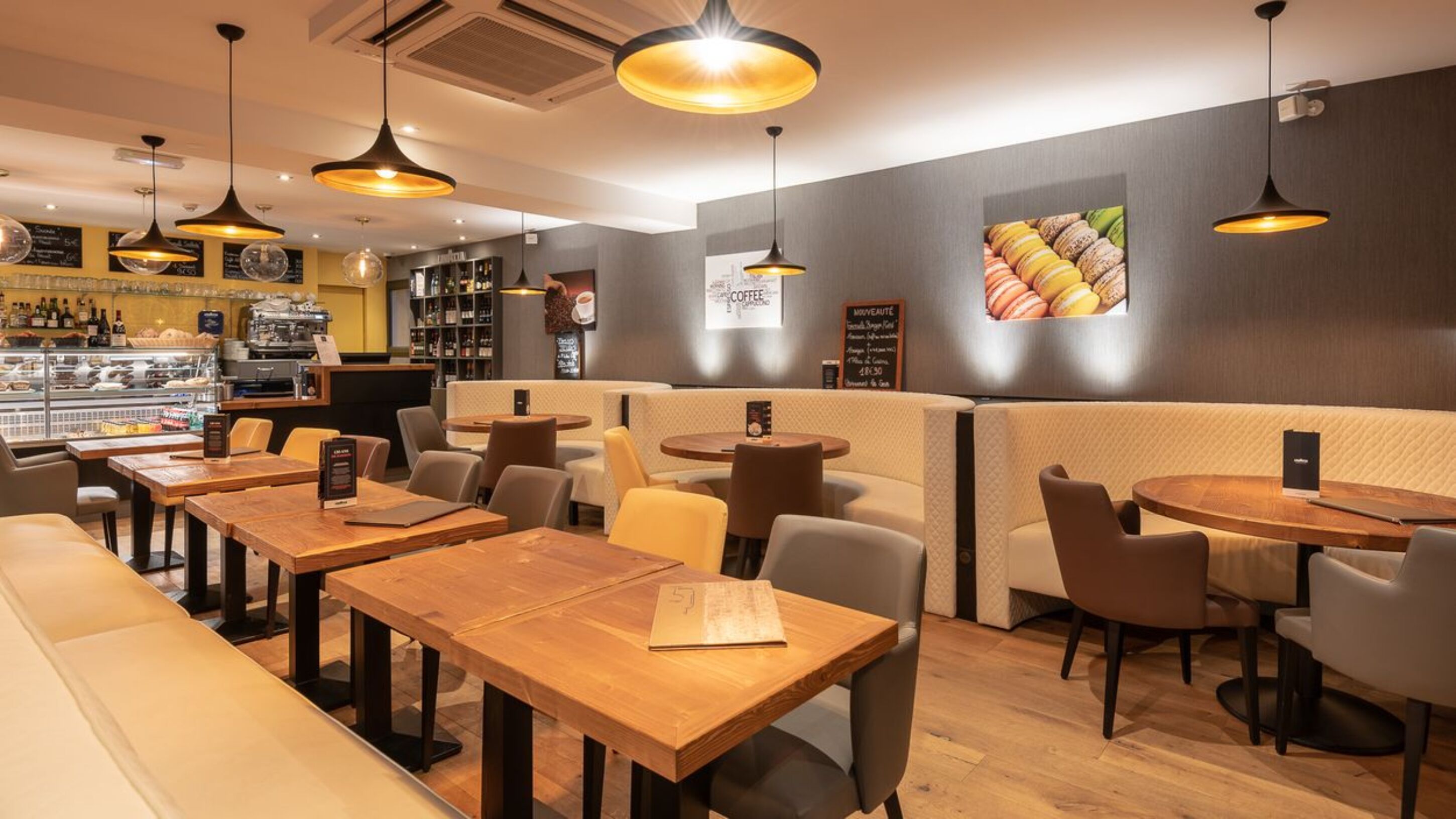 A restaurant interior with wooden tables and chairs, hanging pendant lights, and a wall with a macaron display.