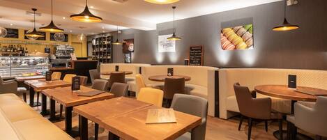 A restaurant interior with wooden tables and chairs, hanging pendant lights, and a wall with a macaron display.
