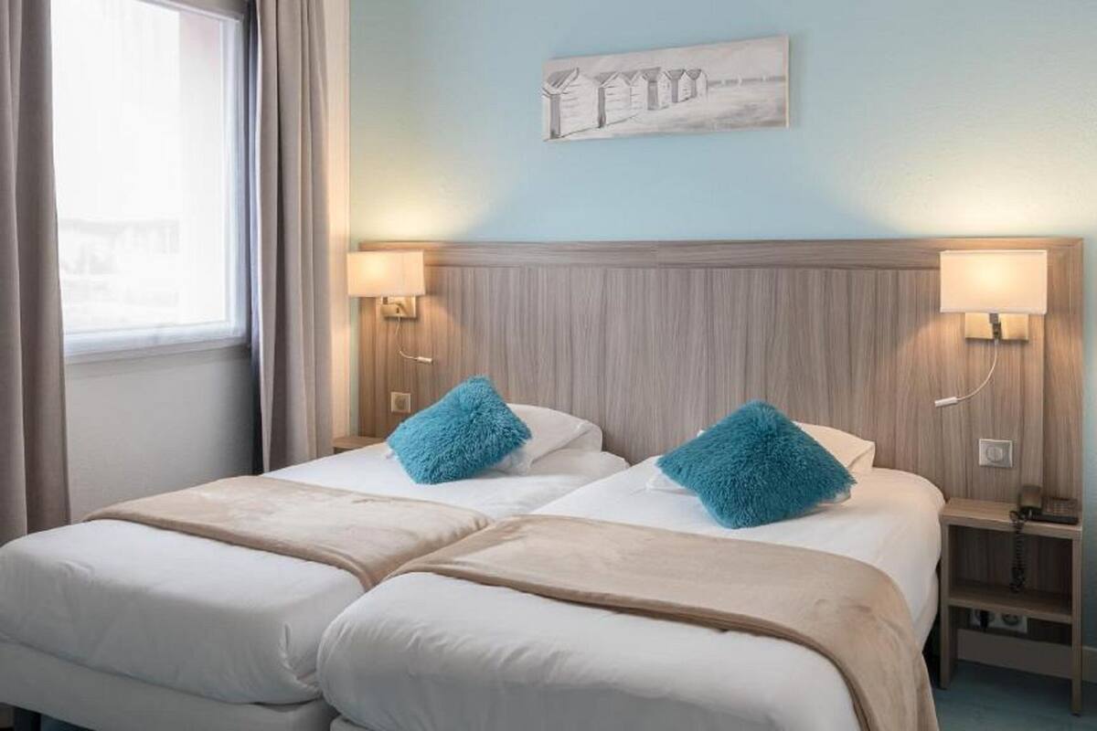 Een hotelkamer met twee bedden, een raam met gordijnen, een kunstwerk aan de muur en bedlampjes.