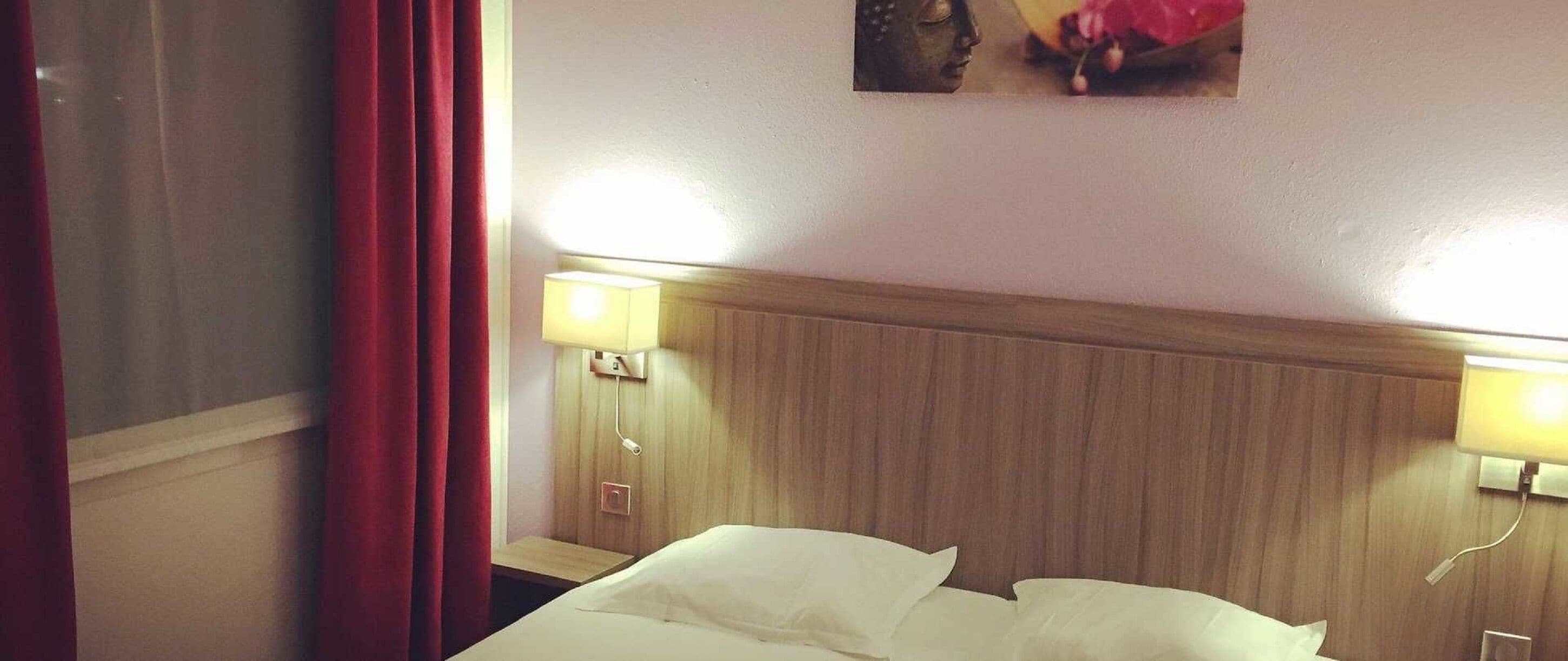 A hotel room with a wooden headboard, two bedside lamps, a bed with white linens, and a wall-mounted artwork.