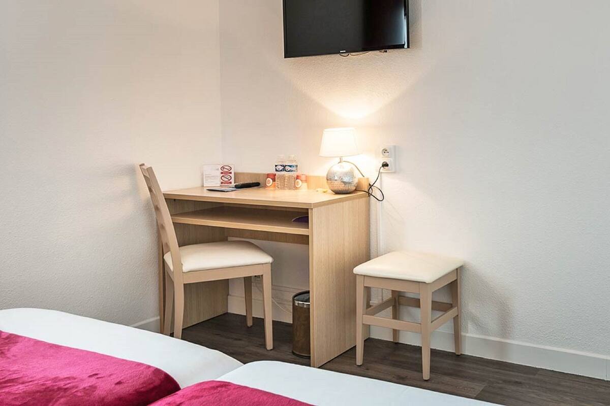 Een hotelkamer met een houten bureau, twee stoelen, een aan de muur bevestigde televisie, een lamp en een prullenbak.