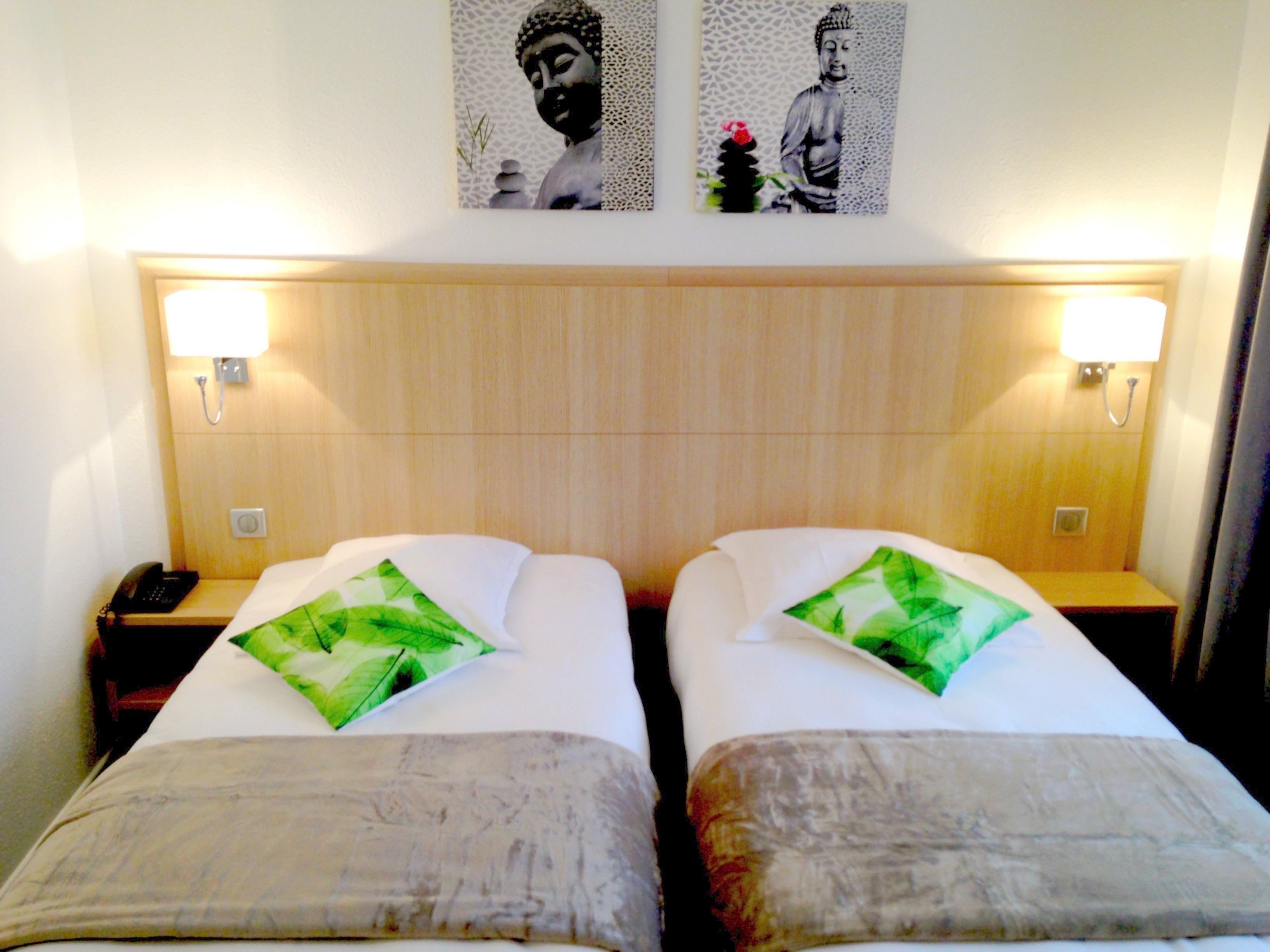A hotel room with two beds, each with a pillow and a bedside table. There are wall-mounted lamps on either side of the beds.