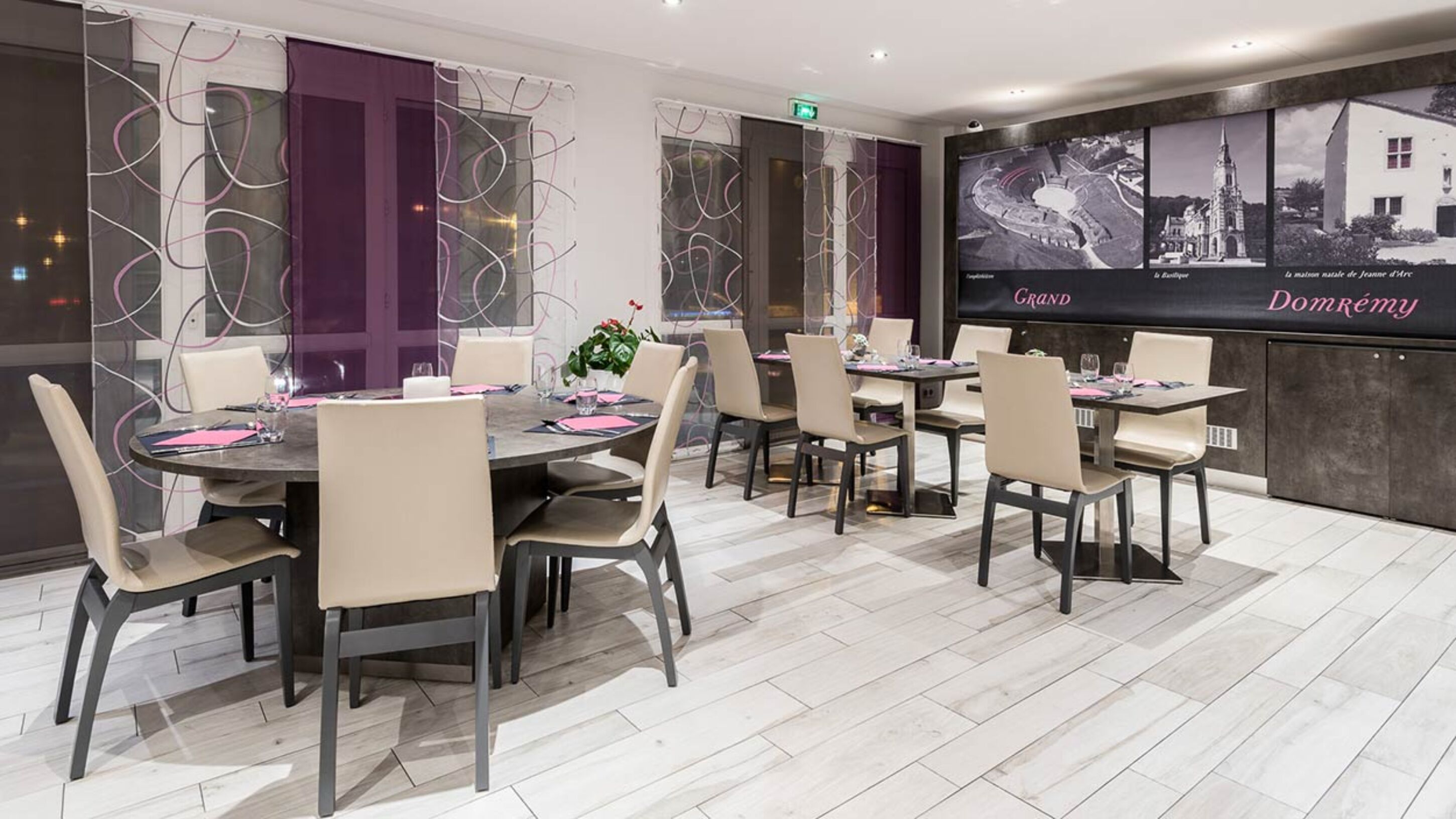 A modern dining area with round tables and chairs, a large wall with historical photos, and a sign reading "Grand Domrémy".