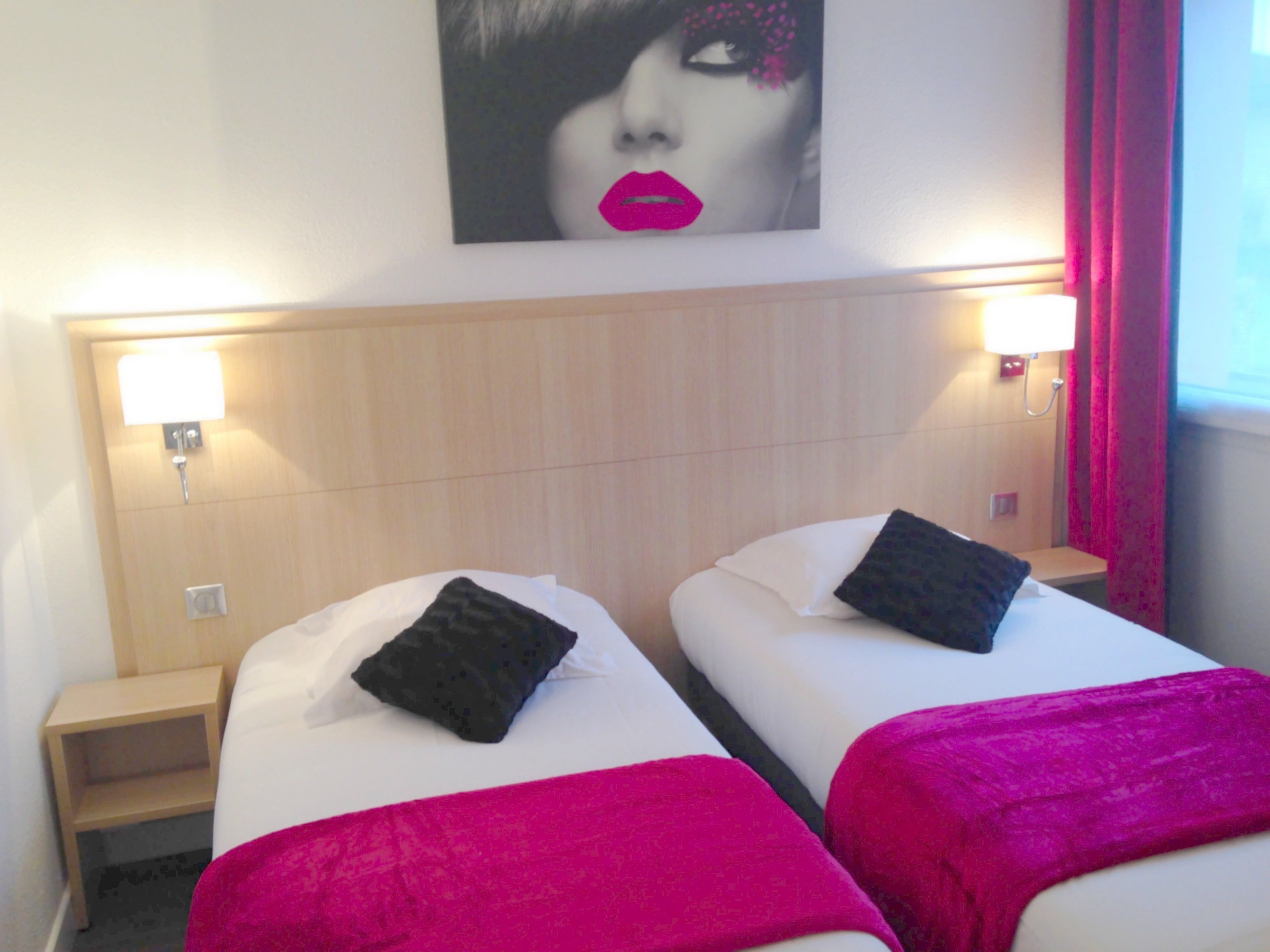 Habitación de hotel con dos camas, cada una con almohada y una manta rosa. En cada lado de las camas hay una mesita de noche con una lámpara.