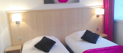 Habitación de hotel con dos camas, cada una con almohada y una manta rosa. En cada lado de las camas hay una mesita de noche con una lámpara.