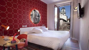Une chambre d’hôtel avec un lit, un miroir rond, une chaise rouge, une table avec une lampe et une vue sur un immeuble par la fenêtre.