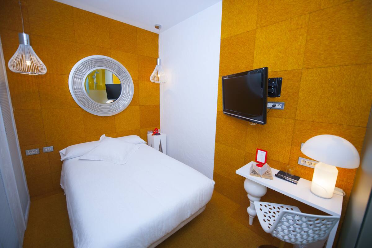 Ein Hotelzimmer mit einem Bett, einem Fernseher, einem Nachttisch mit Lampe, einem Spiegel und einem Wandfernseher.