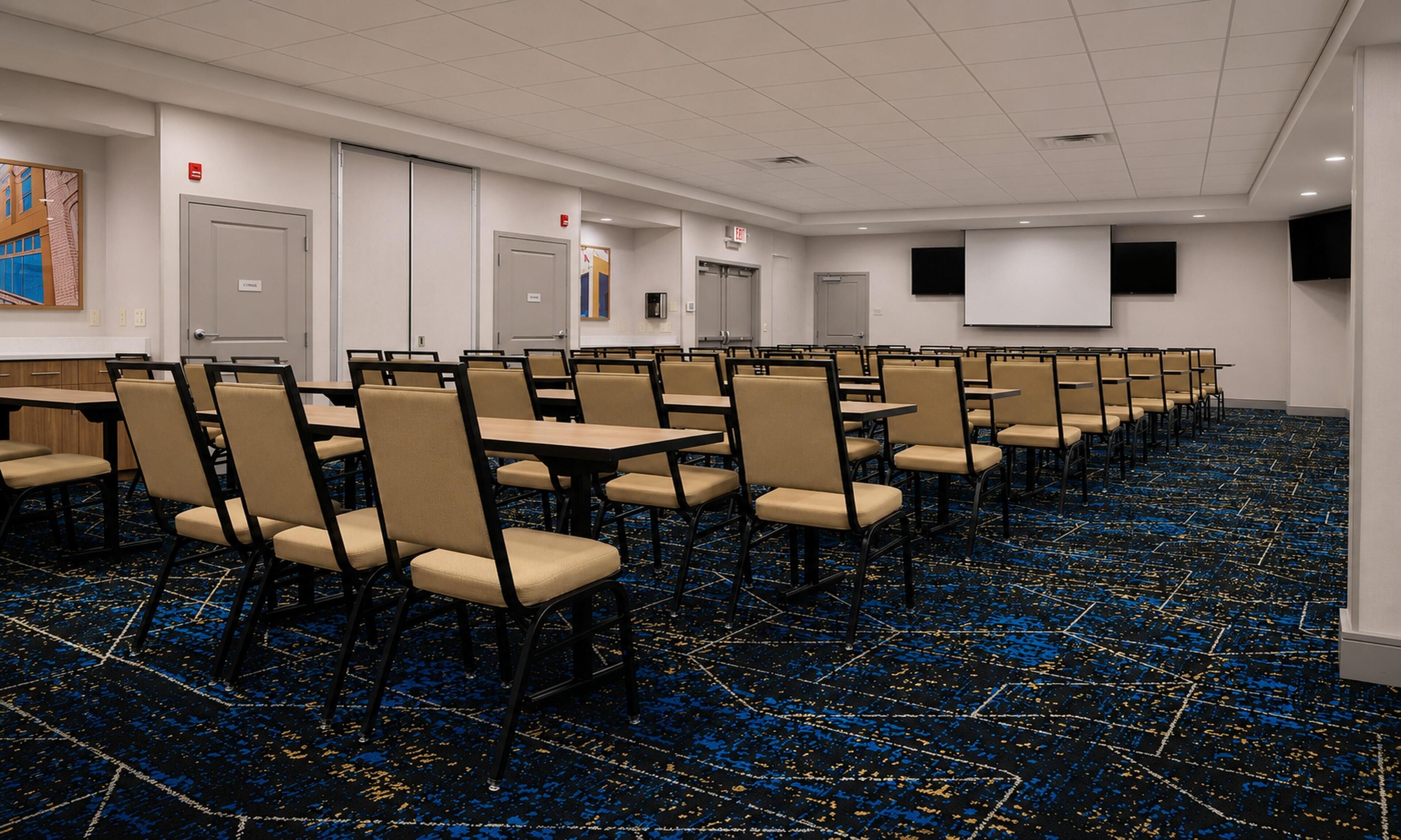 Une salle de conférence avec des rangées de chaises et de tables, un écran de projection et des portes signalées par des panneaux d’indication de sortie.
