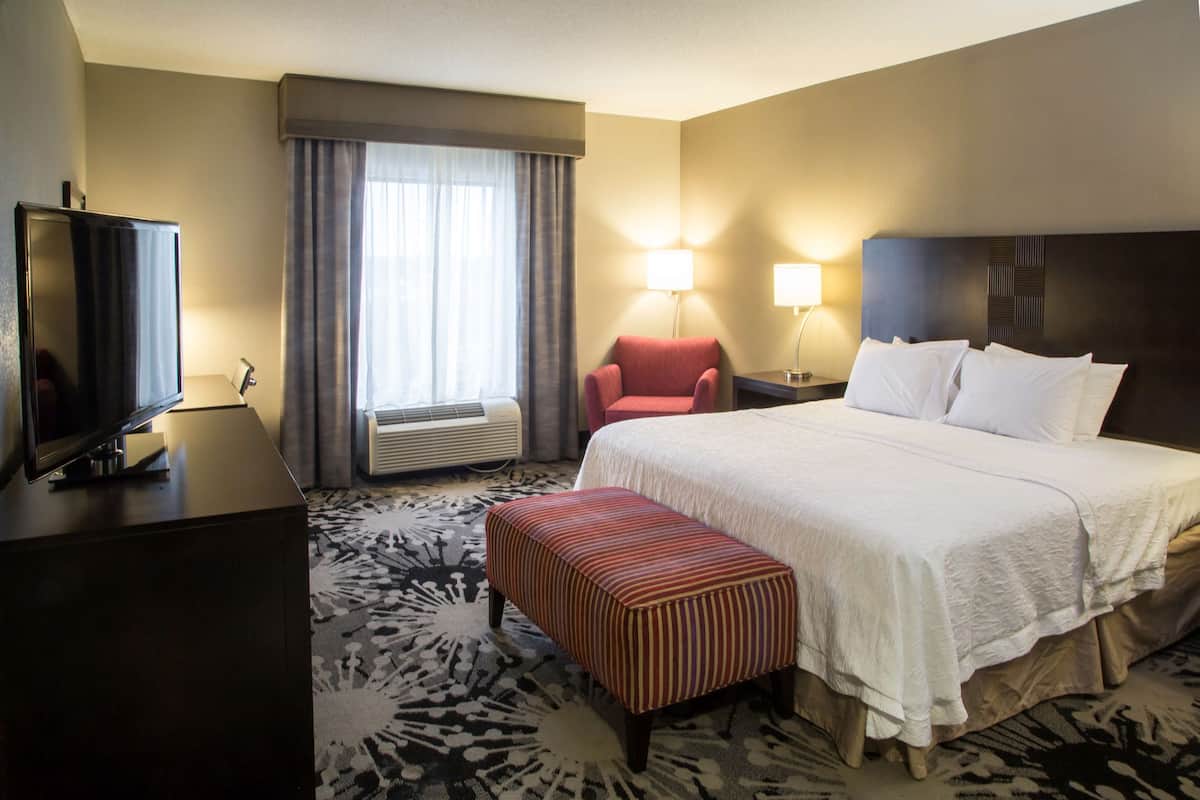 A hotel room with a bed, a television, a chair, a lamp, and an air conditioning unit.