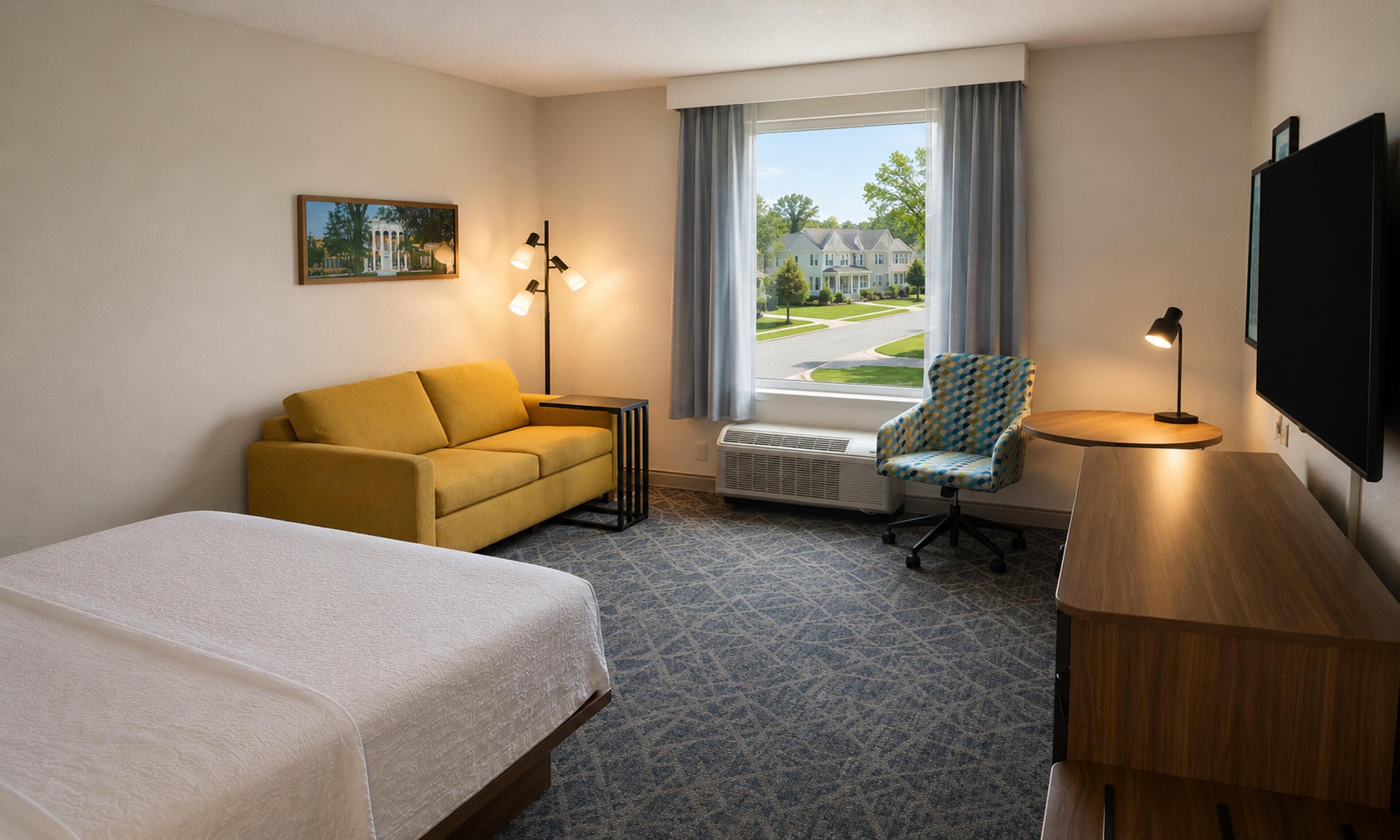 A hotel room with a bed, a sofa, a chair, a desk, a TV, a window with a view, and a picture on the wall.