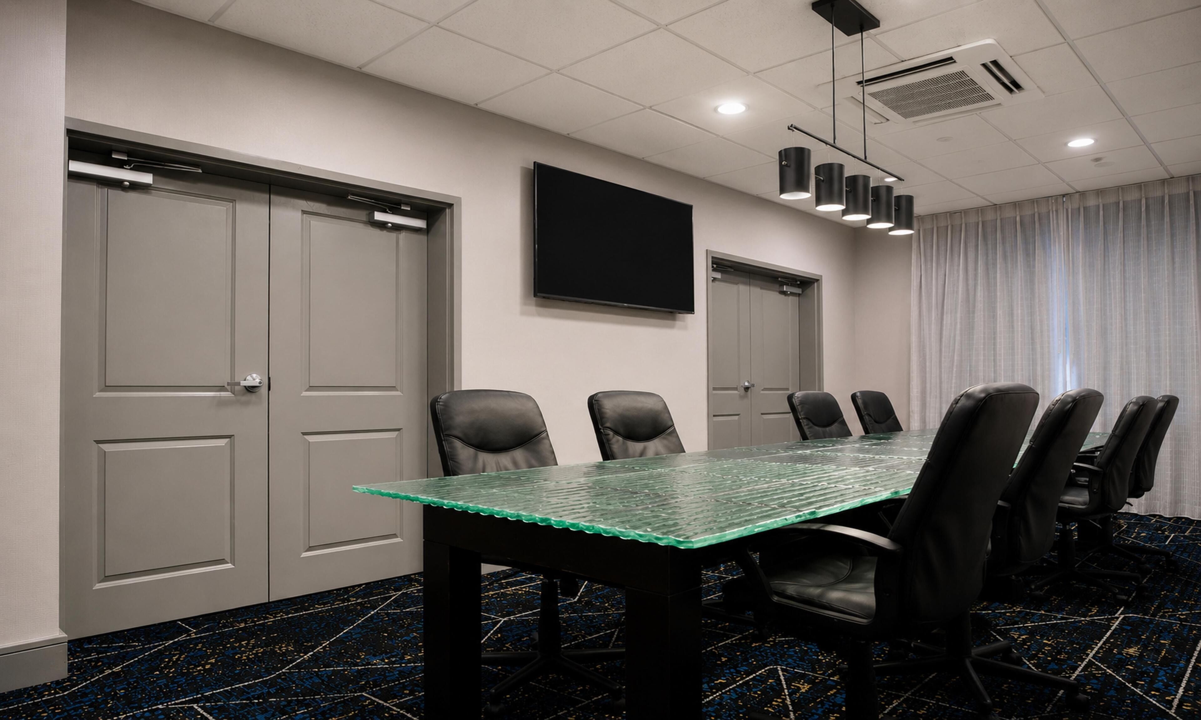 Una sala de conferencias con una mesa de vidrio, sillas negras, un televisor de pantalla plana y una alfombra estampada.