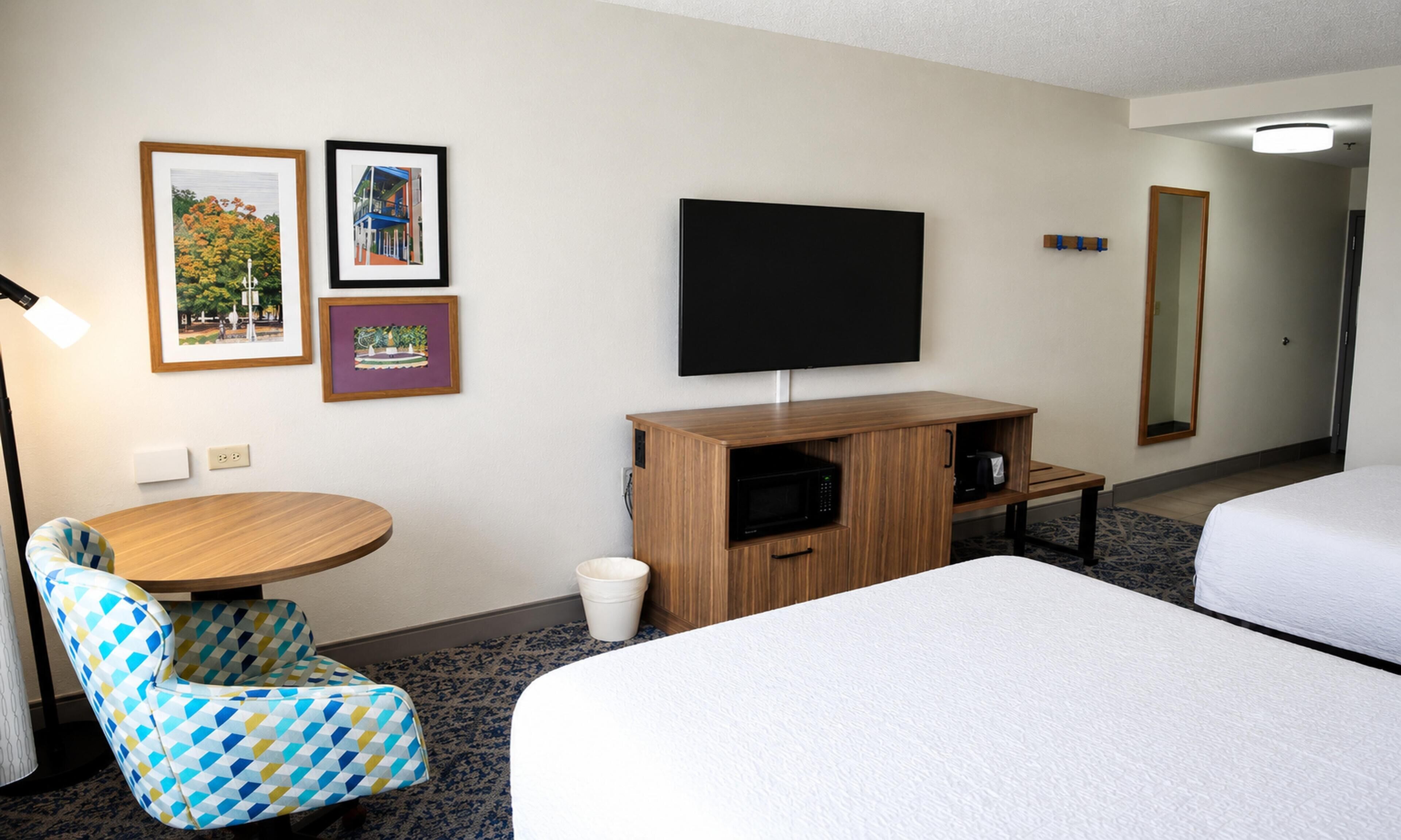 Ett hotellrum med en säng, ett skrivbord, en stol, en tv och inramade tavlor på väggen.