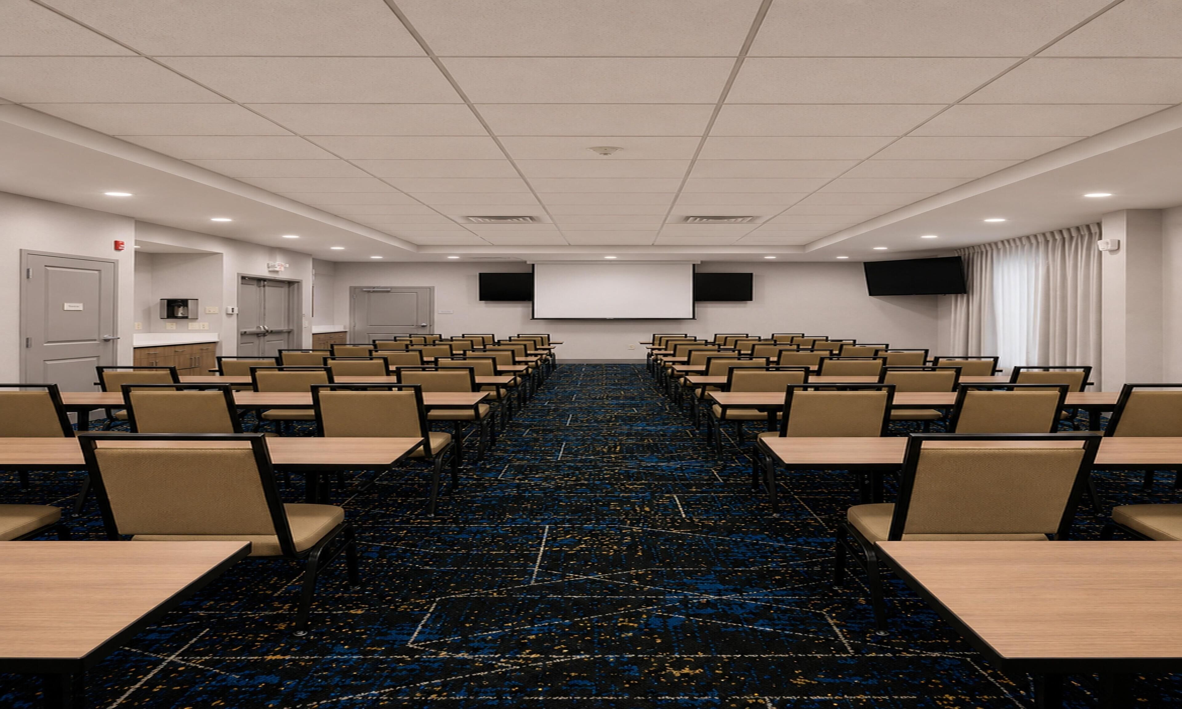 Una sala de conferencias con mesas y sillas dispuestas en filas, una pantalla de proyector y suelo alfombrado.