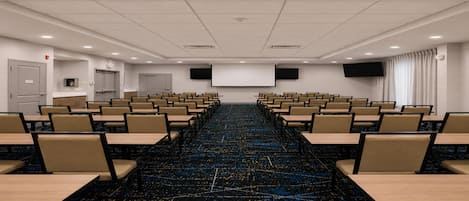 Una sala de conferencias con mesas y sillas dispuestas en filas, una pantalla de proyector y suelo alfombrado.