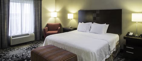A hotel room with a large bed, a chair, a bedside table with a lamp, and a window with curtains.
