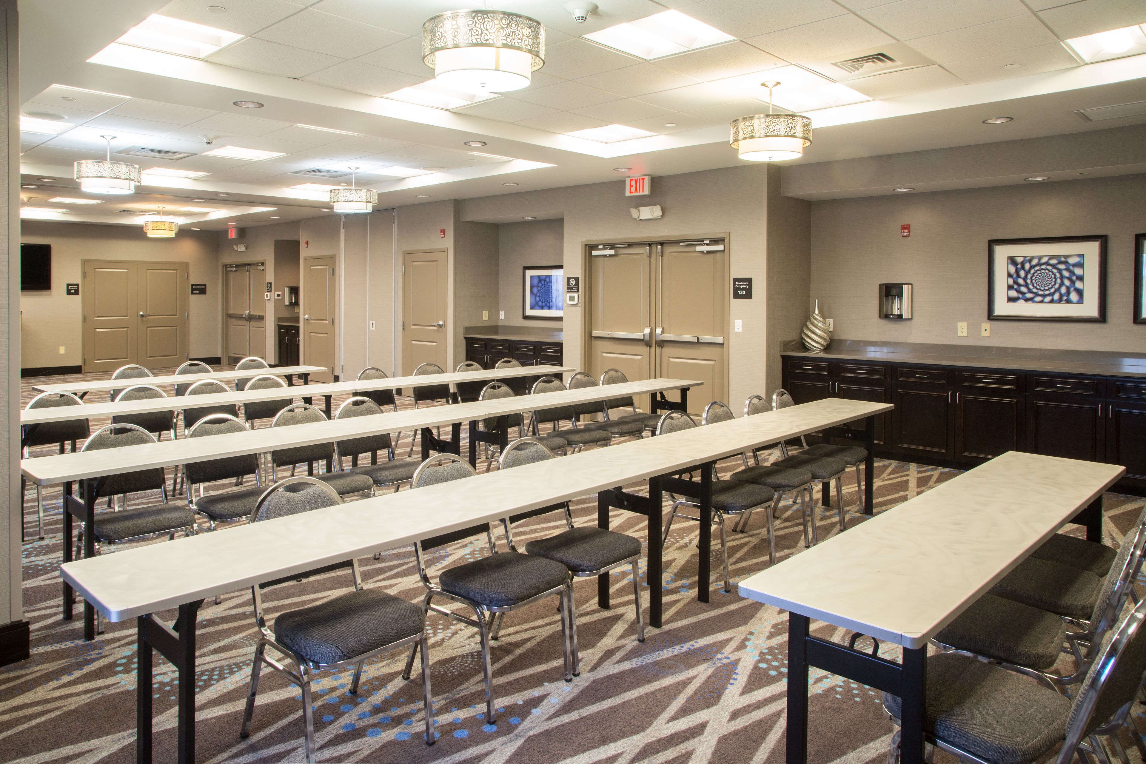 Une salle de conférence dotée de plusieurs tables et chaises, éclairée par des luminaires suspendus et recouverte d’un tapis à motifs.
