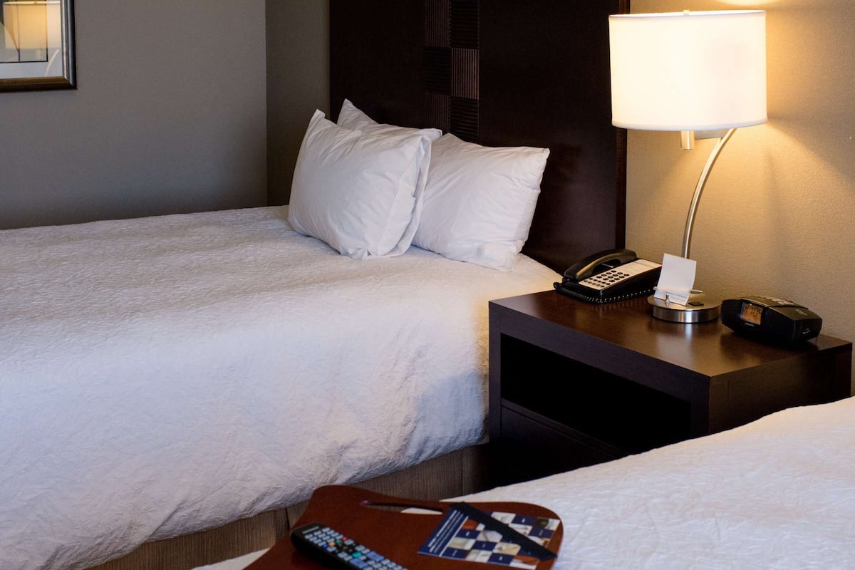 A hotel room with two beds, a bedside table with a lamp, a phone, and a tray table with a remote control.