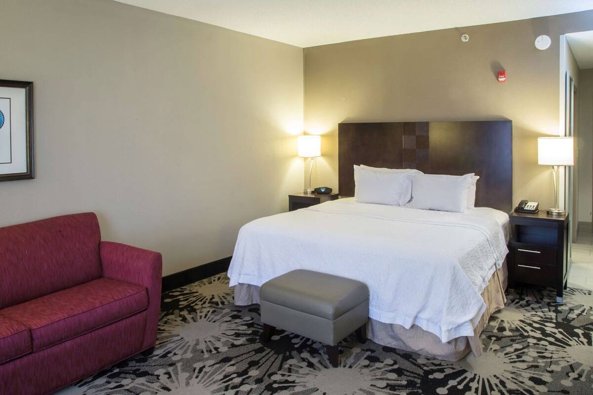 A hotel room with a bed, bedside tables, a red armchair, an ottoman, and a patterned carpet.