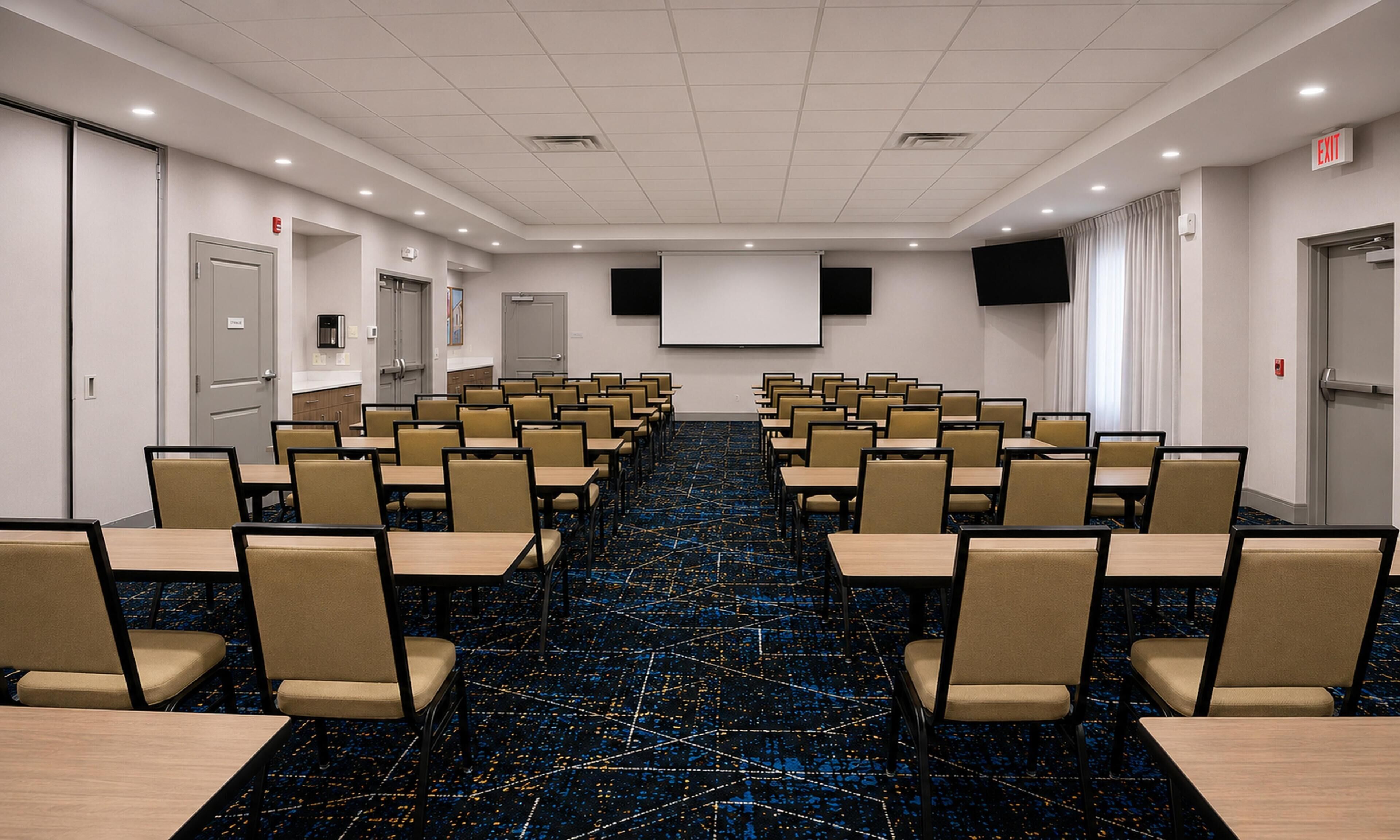 A conference room with rows of tables and chairs, a projector screen, and an exit sign.