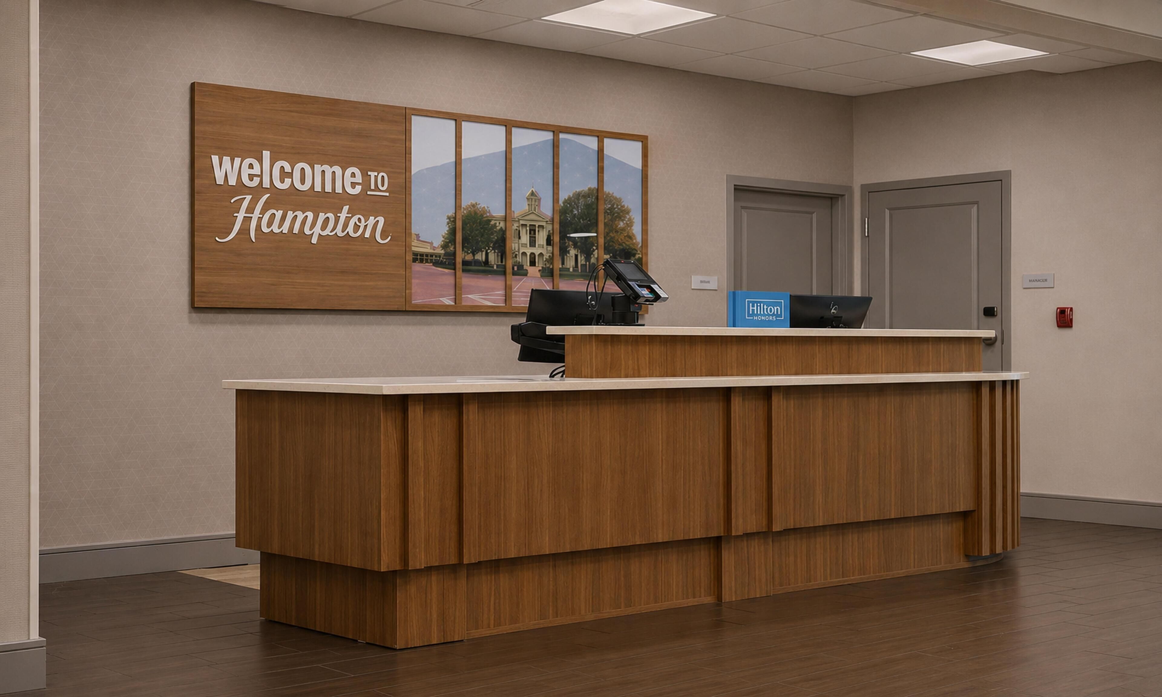 A reception desk with a 'Welcome to Hampton' sign, a computer, and a 'Hilton Honors' box.
