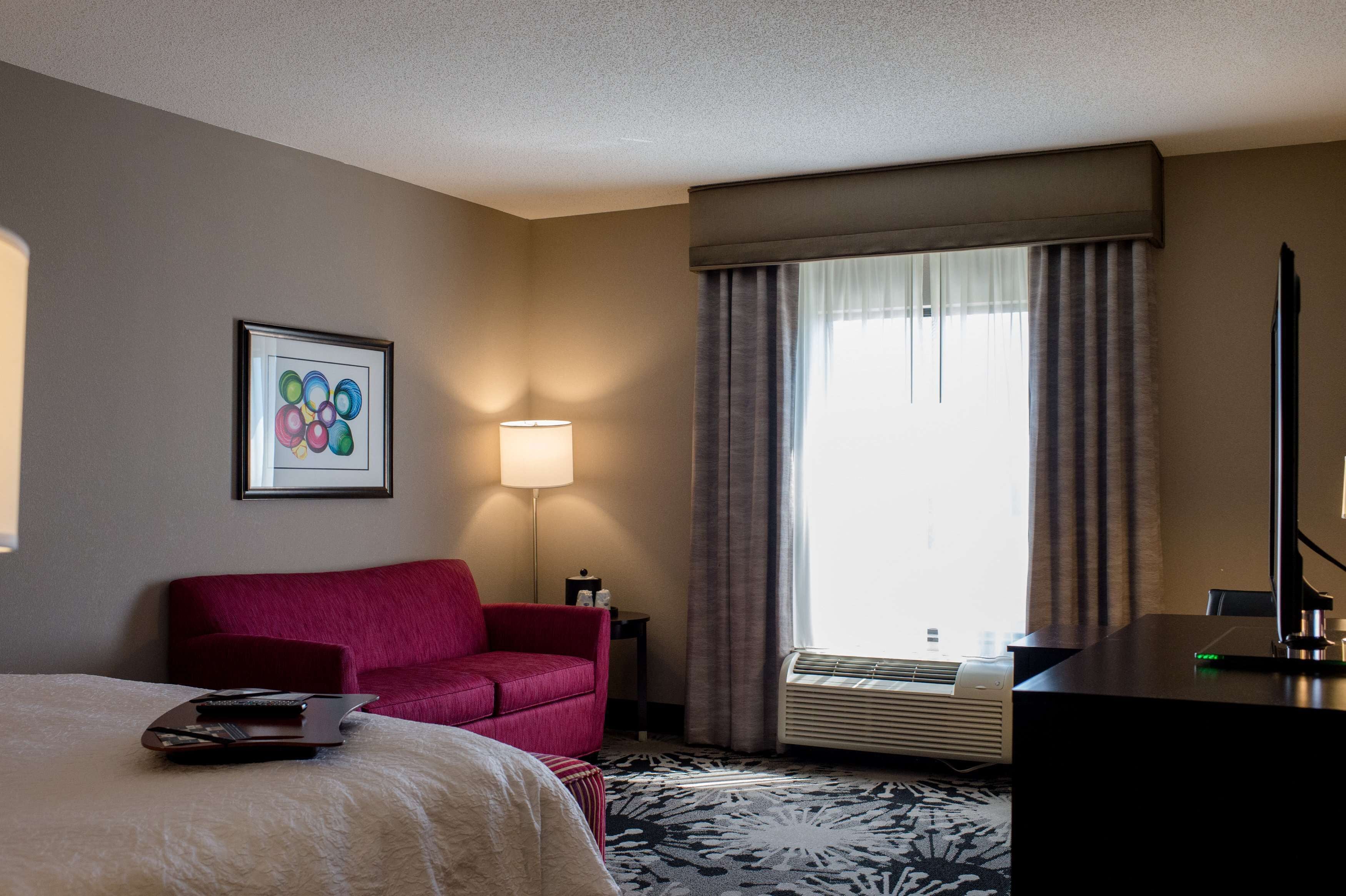A hotel room with a bed, a red sofa, a desk, a lamp, and a large window with curtains.