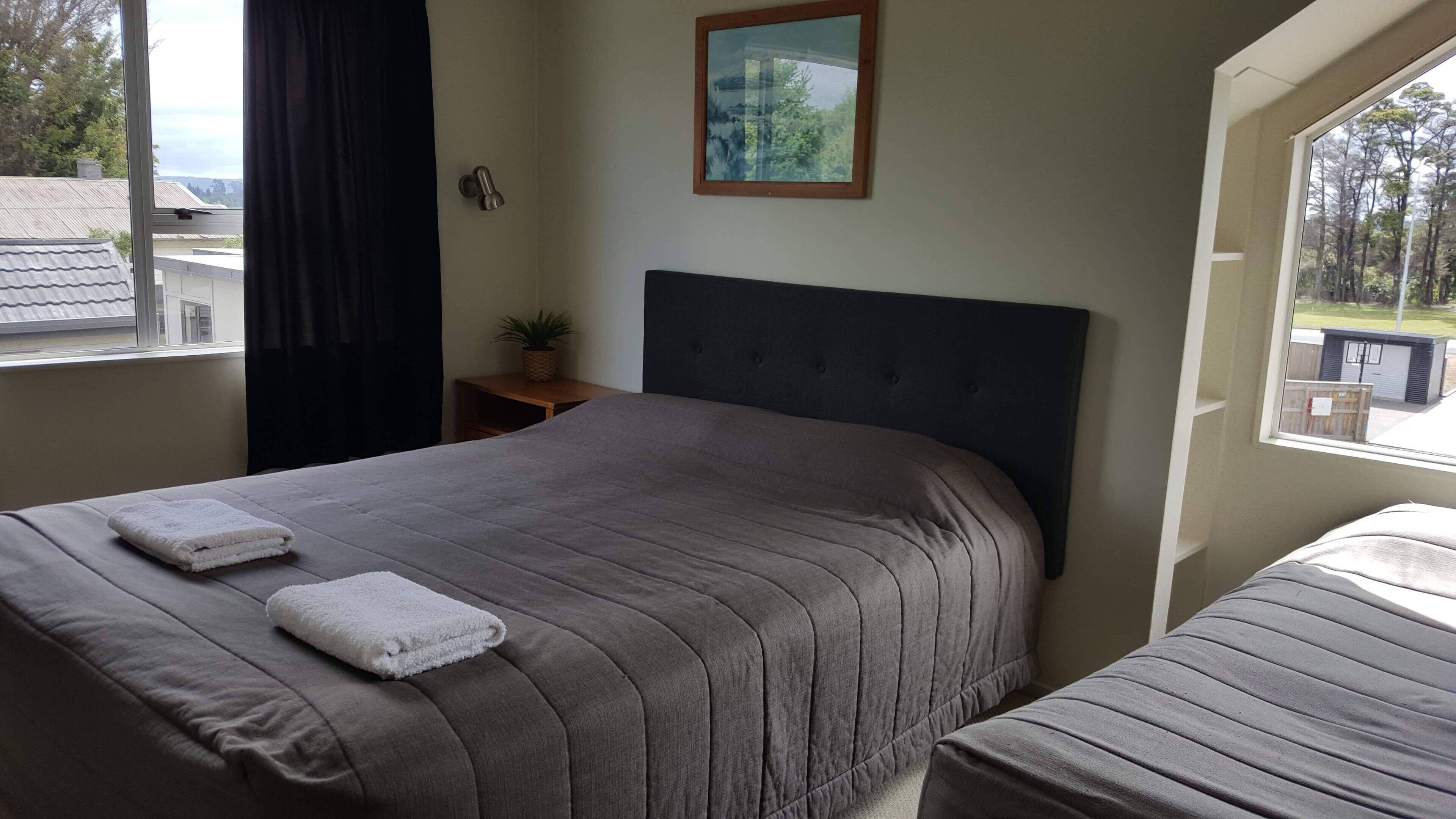Un dormitorio con una cama grande, una cama más pequeña, una ventana con vistas, una planta en una mesita de noche y un cuadro en la pared.