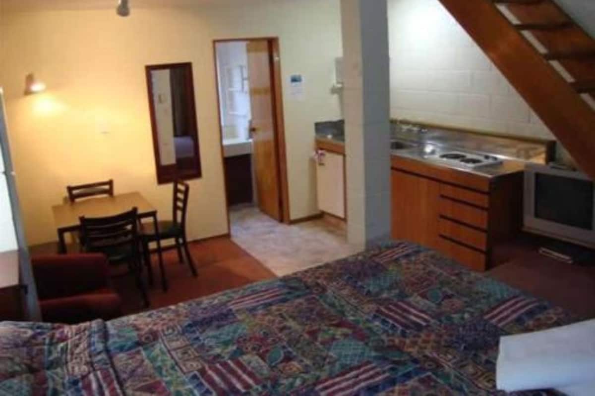 A compact room with a bed, a small dining table, a chair, a kitchenette with a stove and sink, and a mirror.