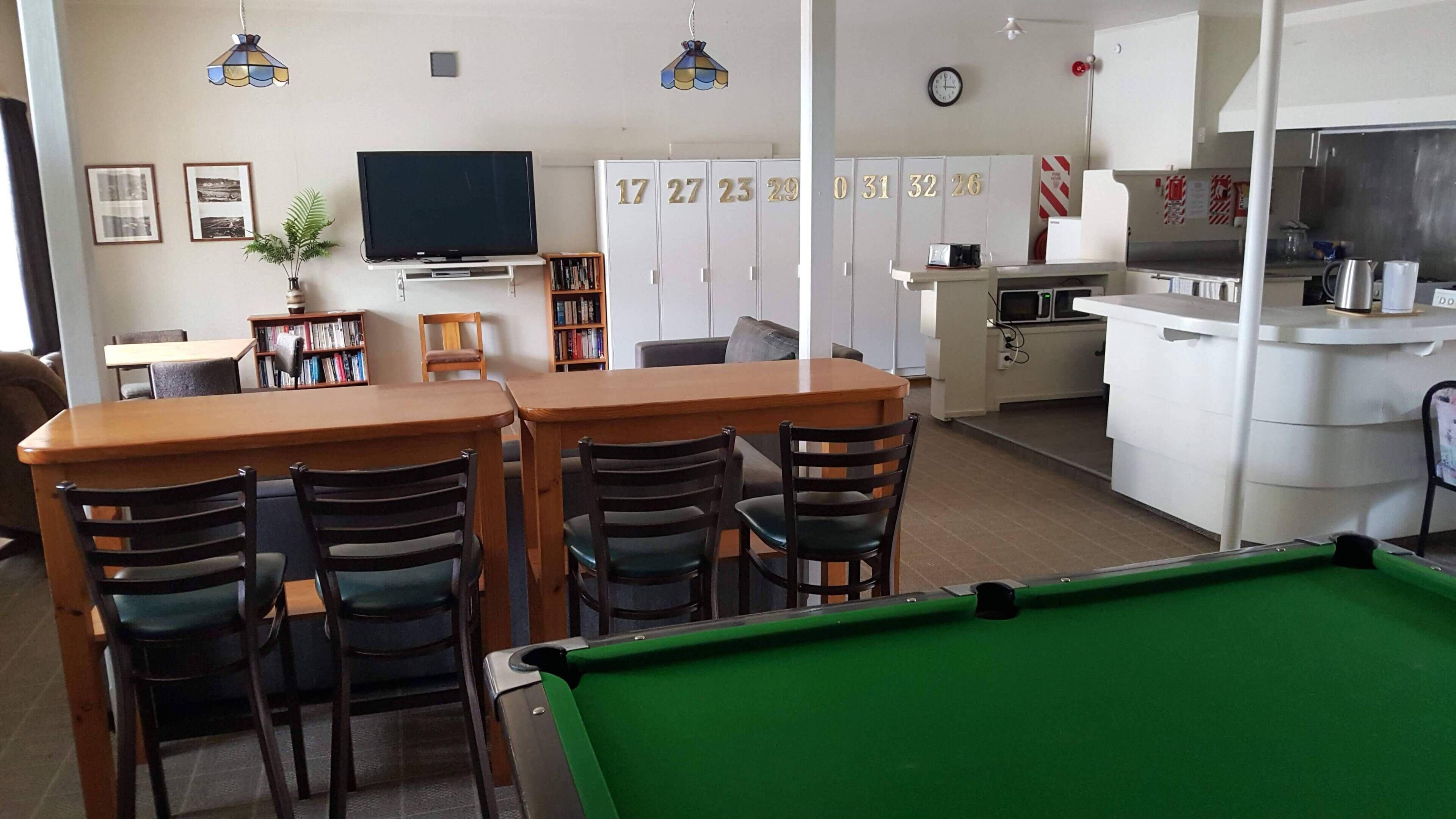 A room with a pool table, a television, a dining area with chairs, a kitchen area, and a set of lockers.