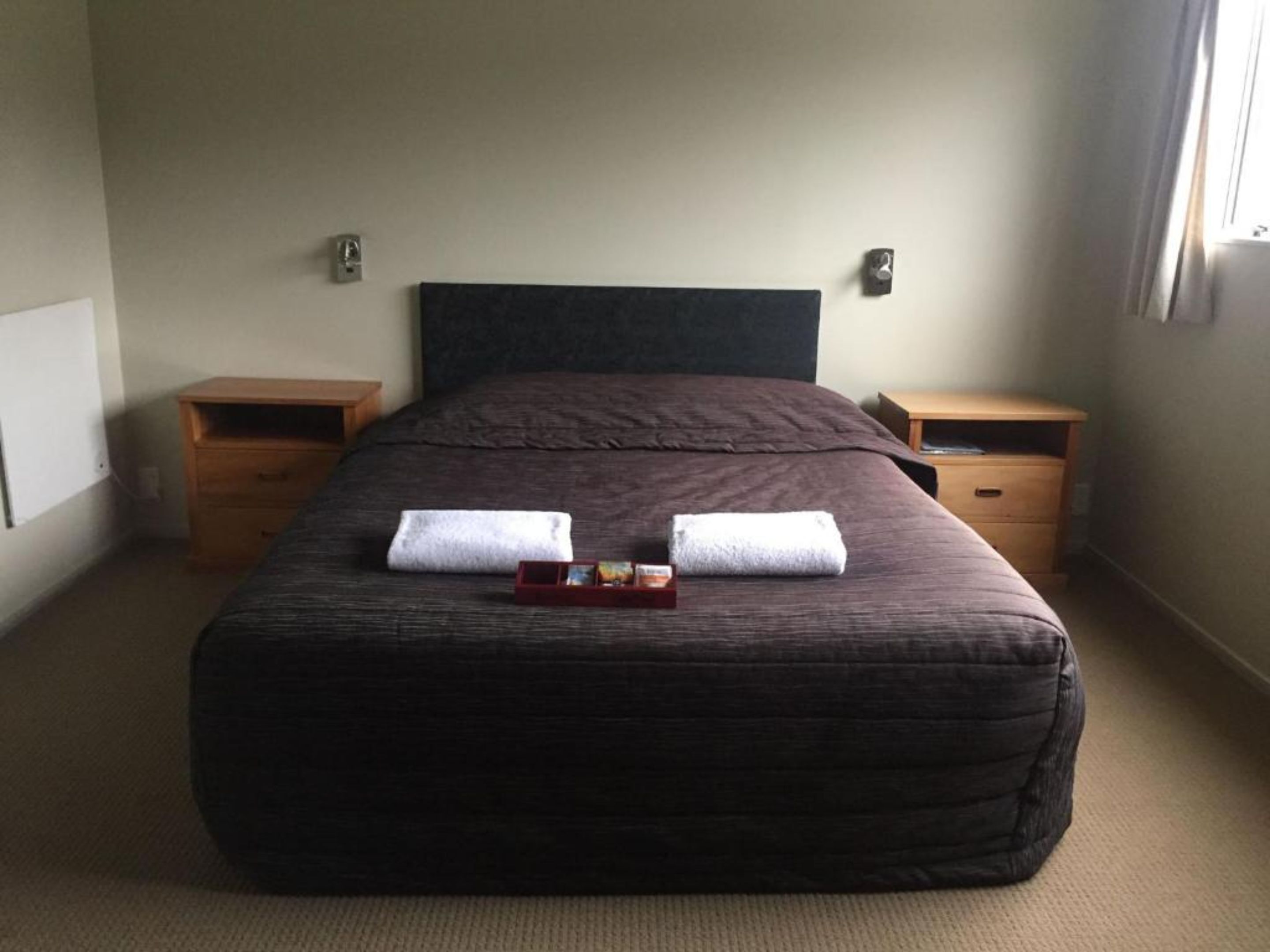 A neatly made bed with two towels, a small tray with a bottle and a glass, and two wooden nightstands.