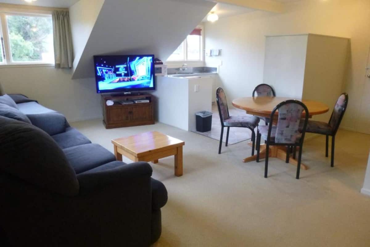 A compact living space with a sofa, a small wooden table, a dining table with chairs, a television, and a kitchenette.