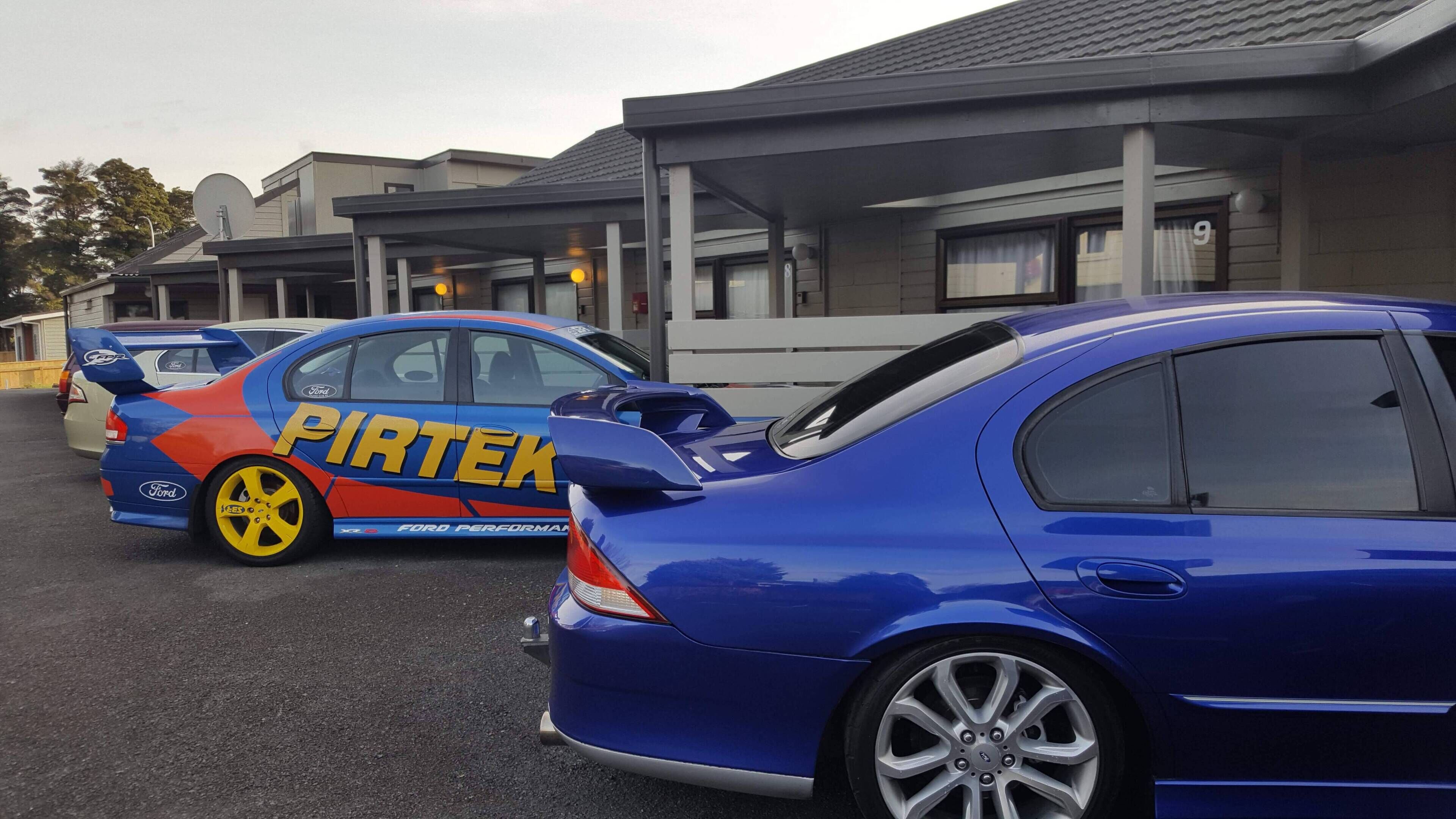 A parking lot with several cars, including a blue sedan and a blue car with 'PIRTEK' branding.