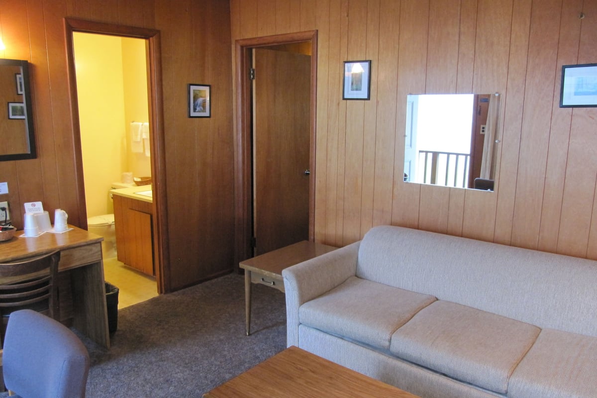 Una habitación con pared de madera, un sofá, una mesa, una silla, un escritorio con teléfono, y un baño visible a través de una puerta abierta.