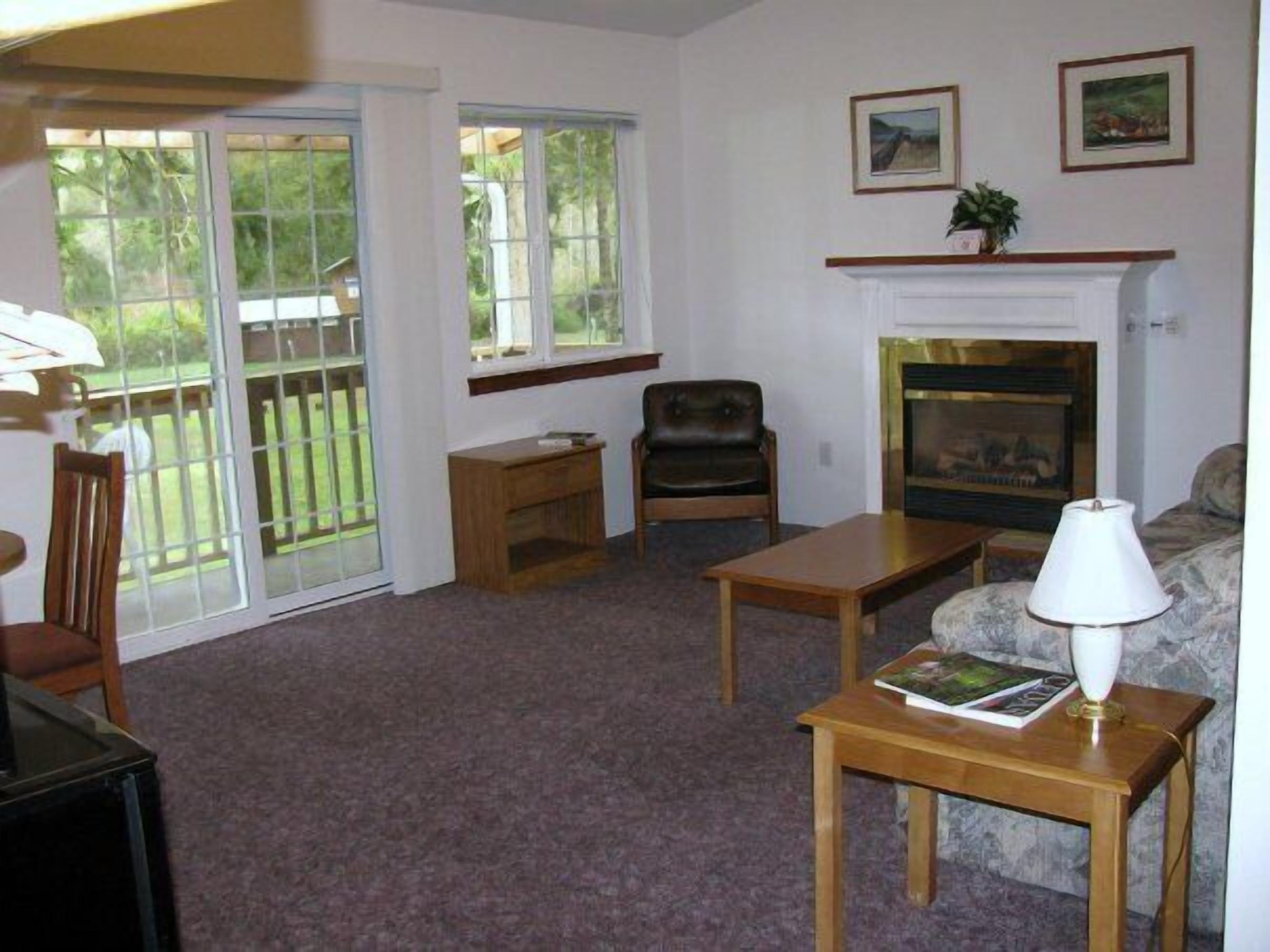 A living room with a fireplace, a sofa, a coffee table, and a chair. There are two windows and a door leading to a balcony.