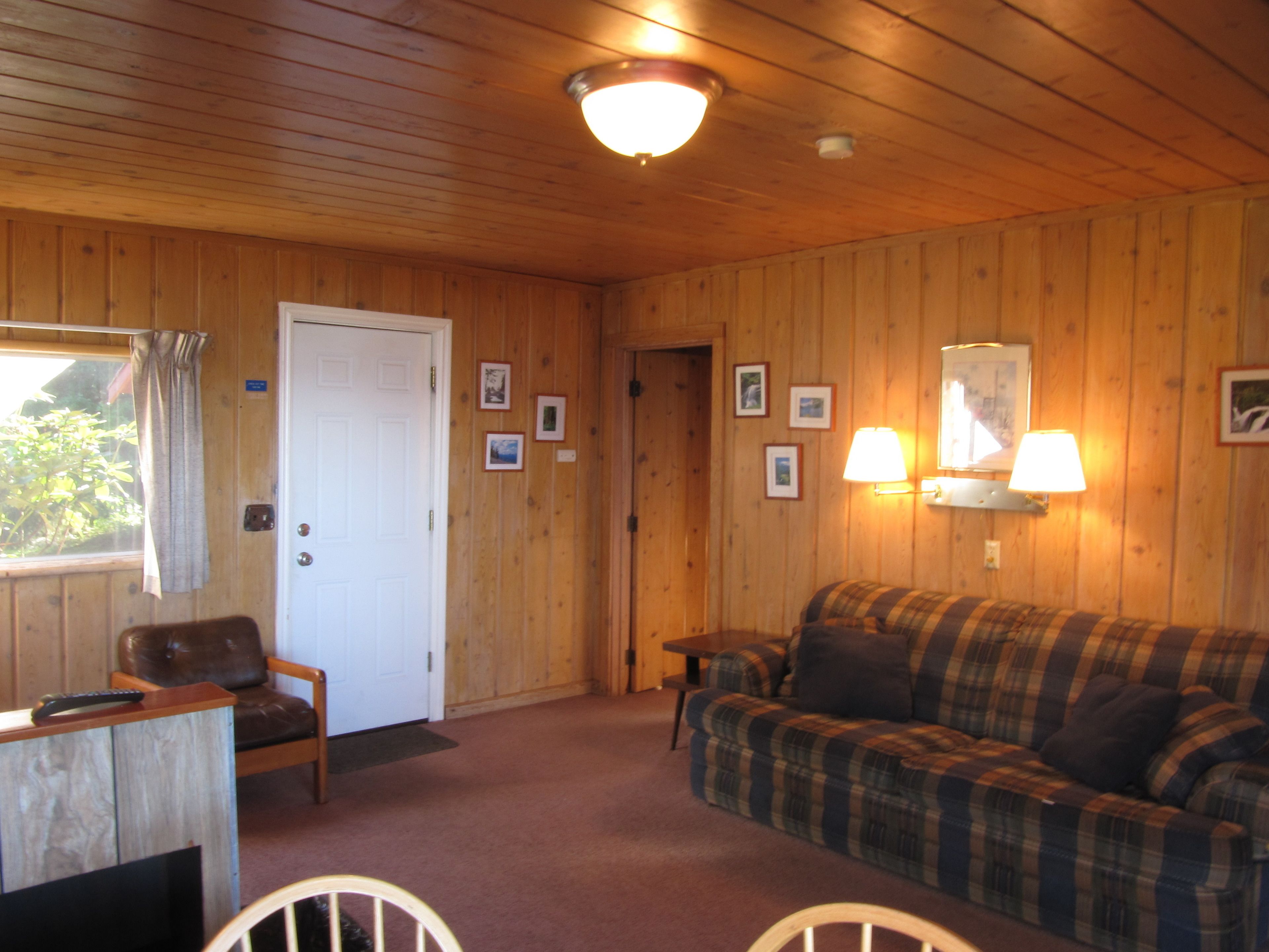 A cozy room with wooden walls, a plaid sofa, a chair, a desk with a remote control, and framed pictures on the wall.