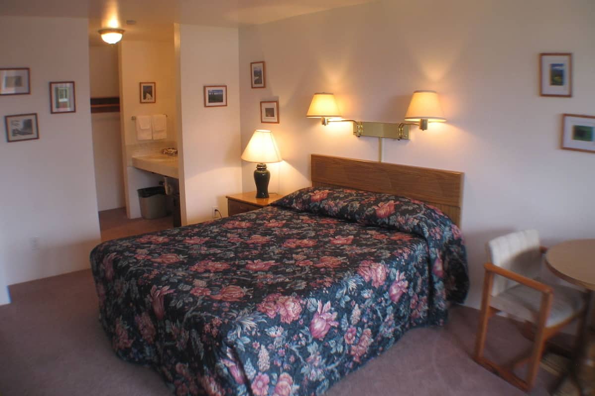 Habitación de hotel con cama, dos lámparas de noche, una mesita, una silla y un baño a la vista al fondo.