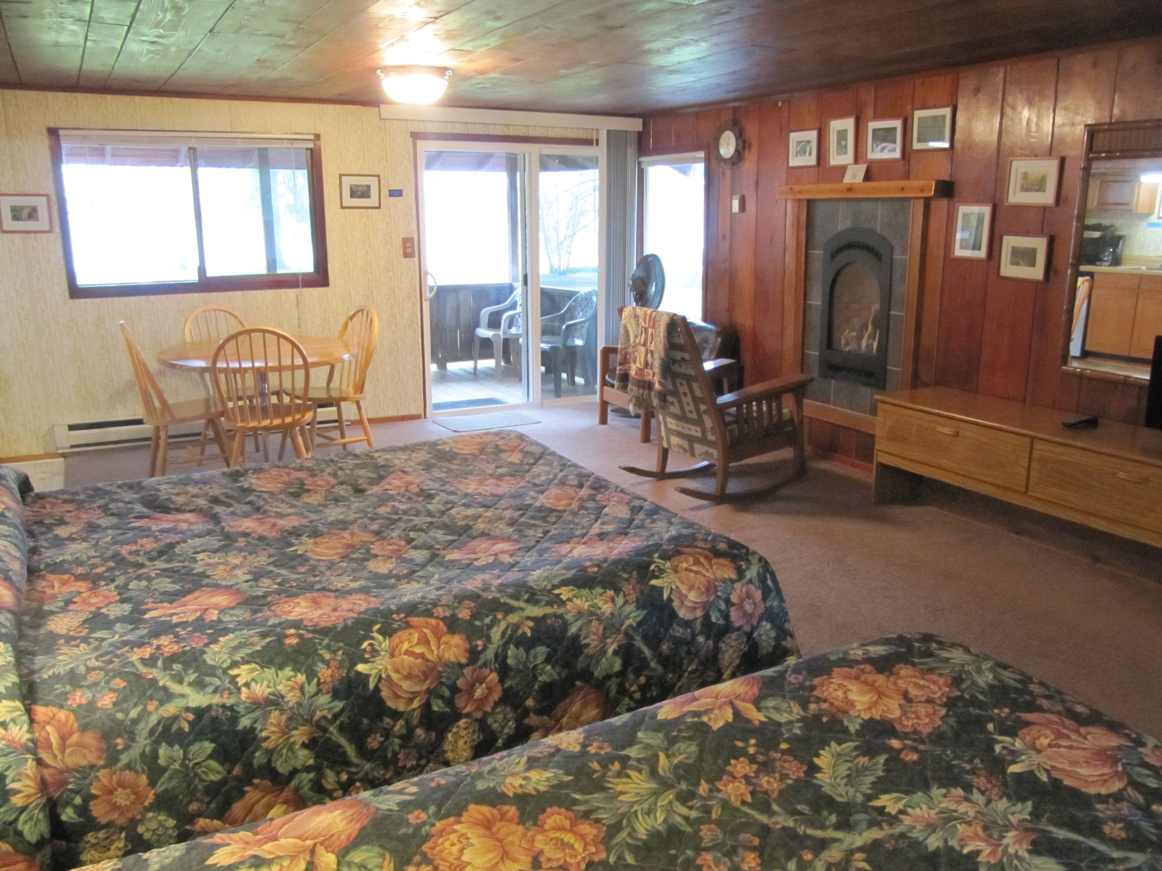 A room with two beds, a fireplace, a dining area with chairs, and a view through a glass door.
