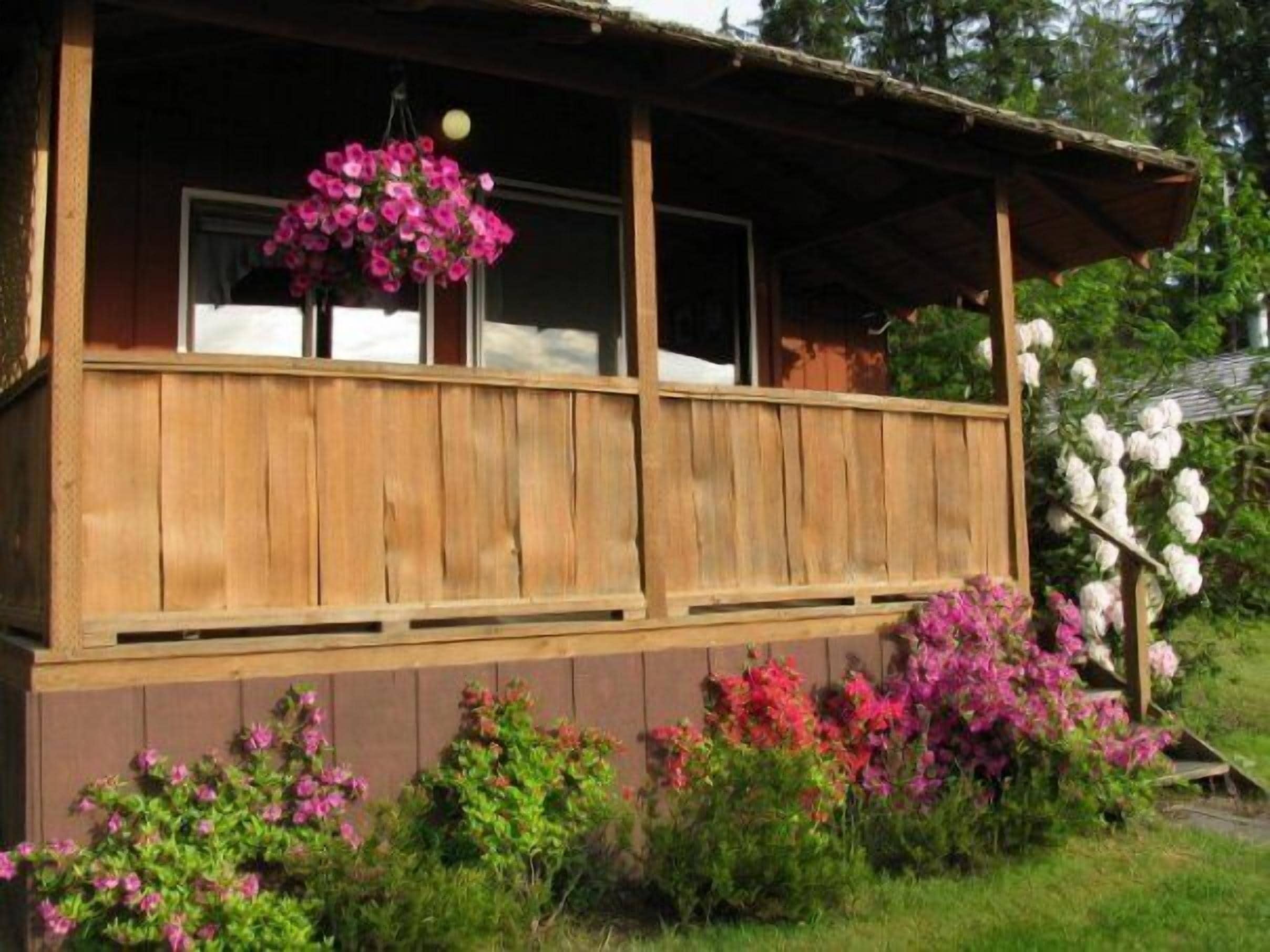 Une cabane en bois dotée d’un porche, entourée de plantes en fleurs.