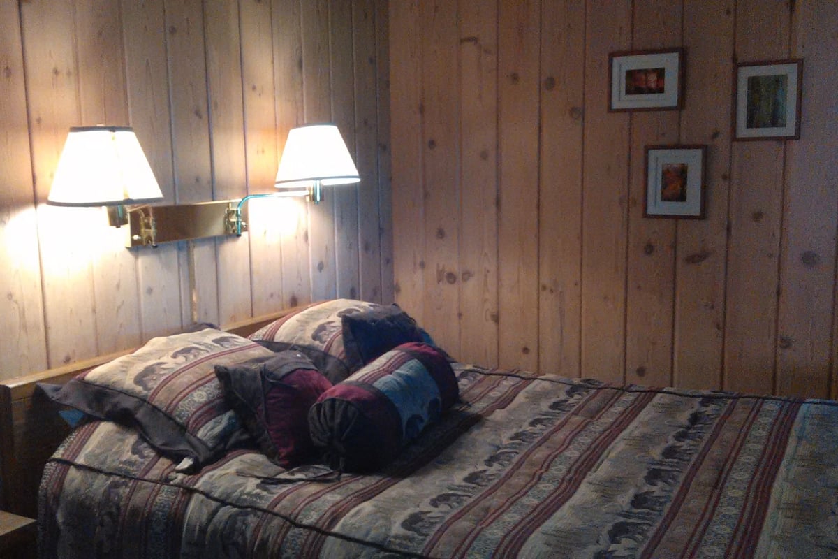 Un dormitorio con pared de madera, dos lámparas de noche, una cama con ropa de cama estampada y tres cuadros en la pared.