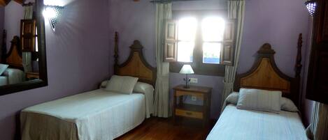 A room with two single beds, wooden beams on the ceiling, a small bedside table with a lamp, and a window with curtains.