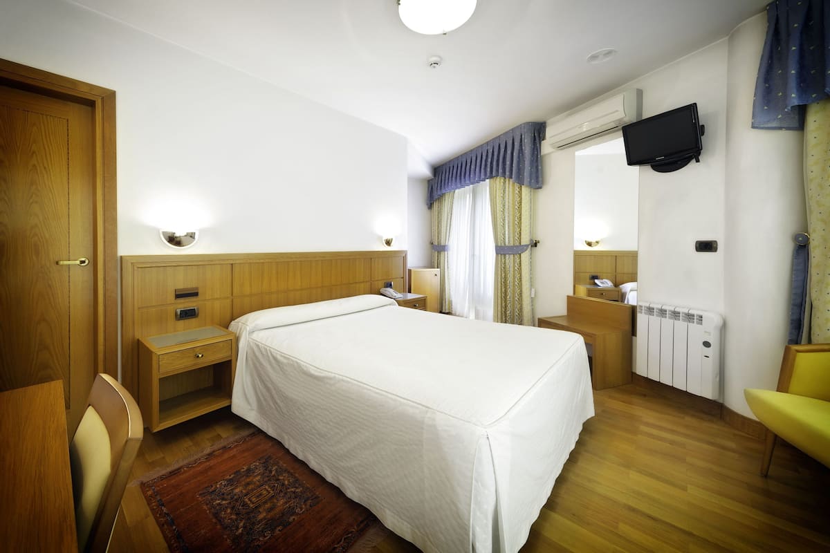 Ein Hotelzimmer mit Bett, Nachttischen, Schreibtisch, Stuhl, an der Wand montiertem Fernseher und Heizkörper.