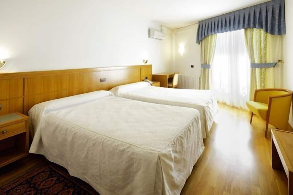 Ein Hotelzimmer mit zwei Betten, einem hölzernen Kopfteil, einem Nachttisch, einem Stuhl, einem Schreibtisch und einem Fenster mit Vorhängen.