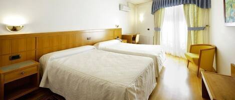 A hotel room with two beds, a wooden headboard, a bedside table, a chair, a desk, and a window with curtains.