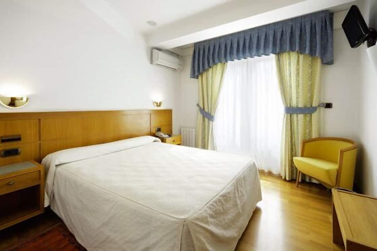 Ein Hotelzimmer mit hölzernem Kopfteil, einem Bett mit weißer Bettdecke, einem Nachttisch mit Lampe, einem Sessel und einem Fenster mit Vorhängen.