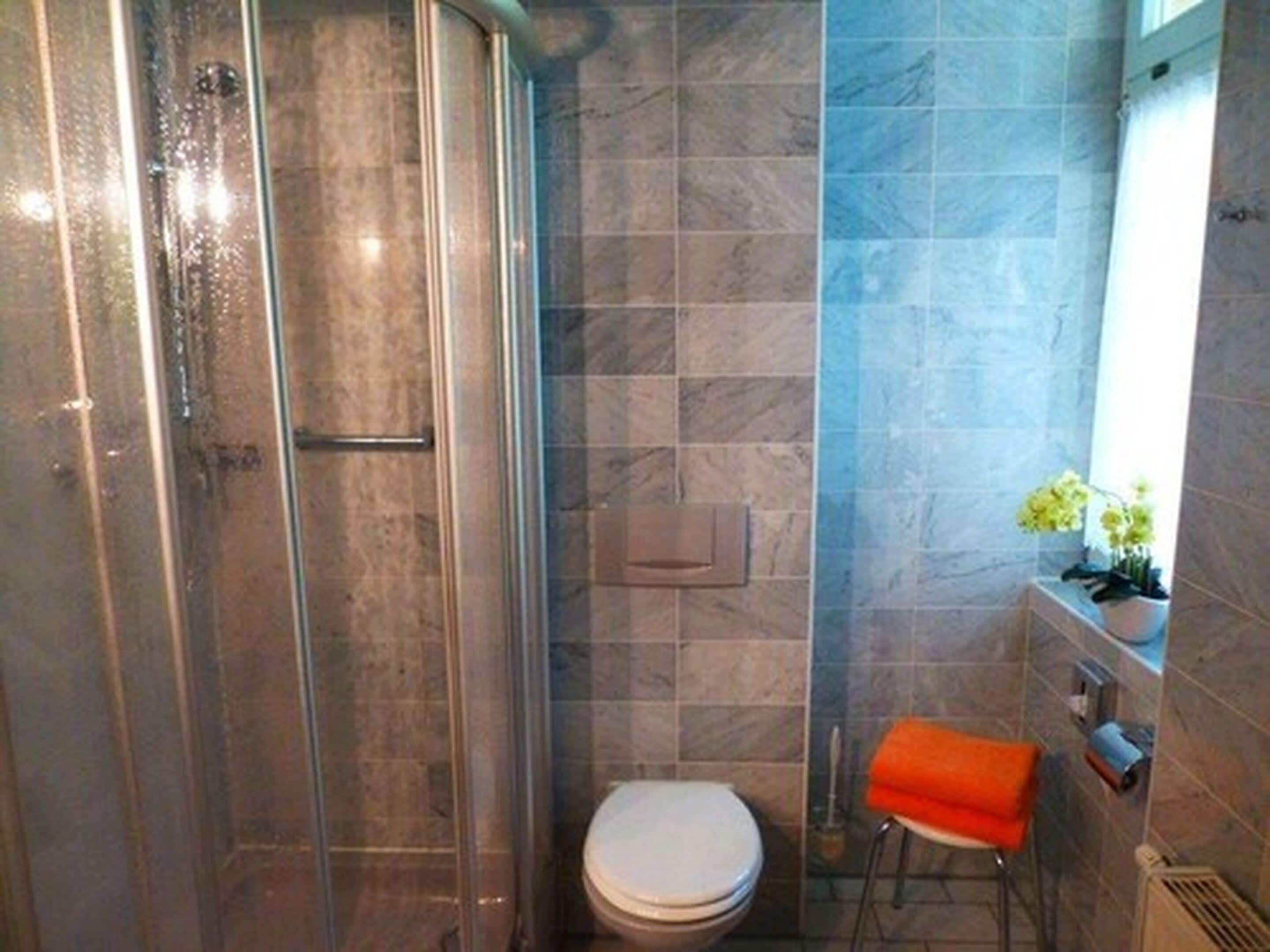 A bathroom with a glass-enclosed shower, a toilet, and a small stool with an orange towel.