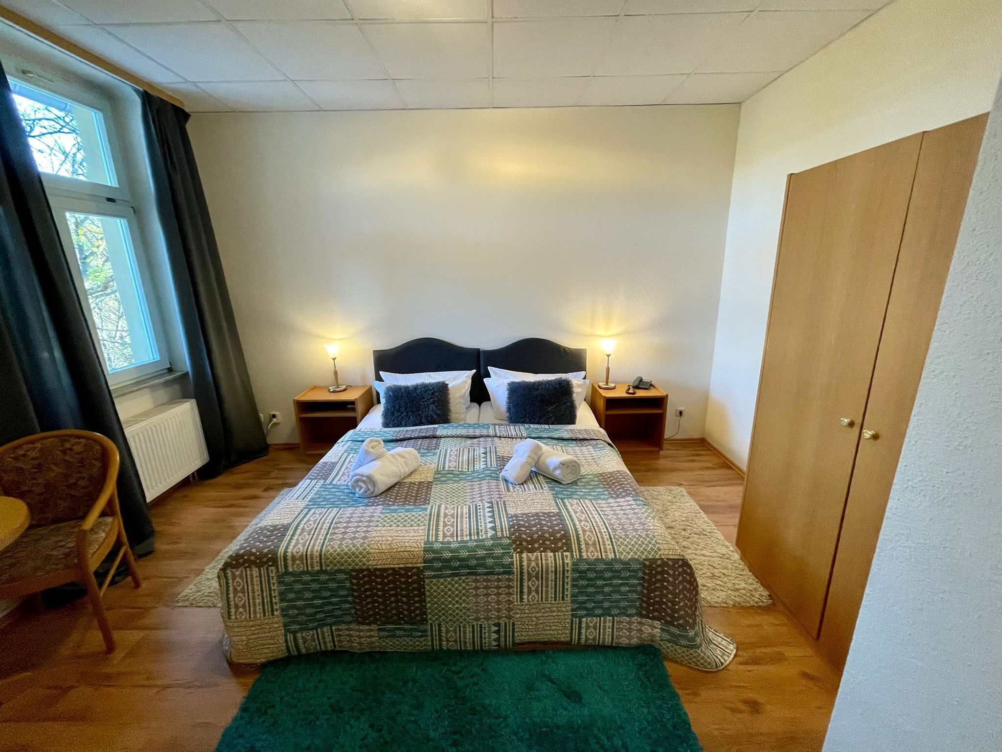 Una cama bien hecha con una colcha estampada, dos lámparas de noche, una silla y una ventana con cortinas.