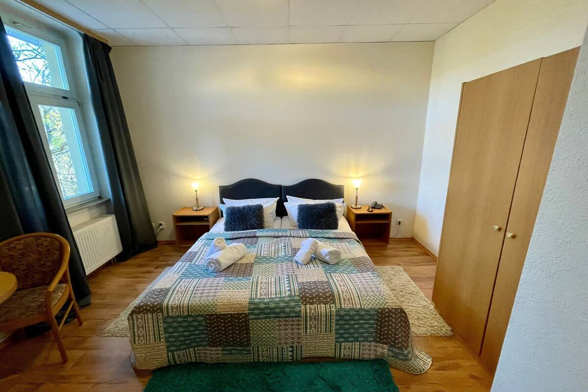 A neatly made bed with a patterned bedspread, two bedside lamps, a chair, and a window with curtains.