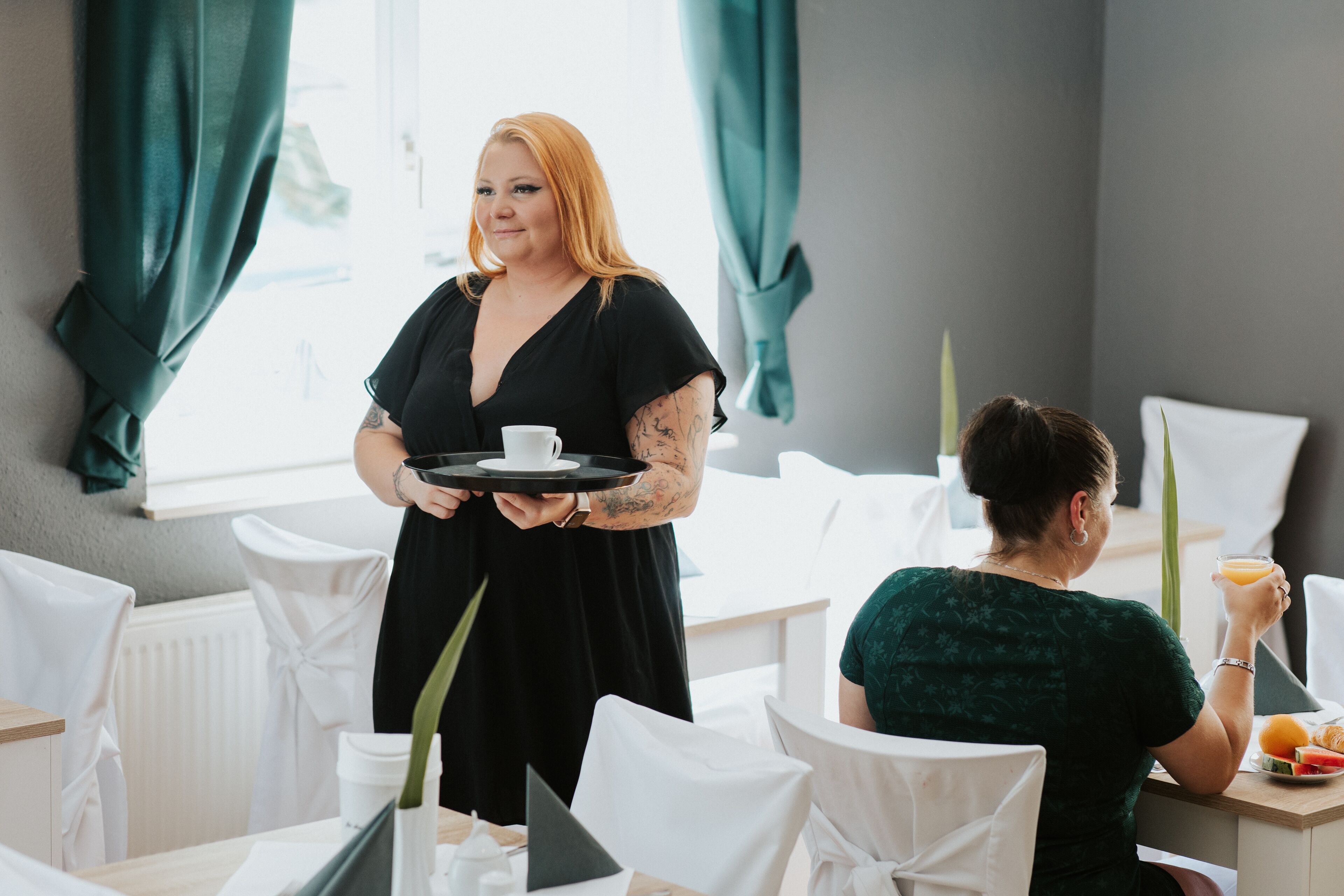Hotellin ruokailutila, jossa on valkoisia tuoleja, pöytä, jonka päällä on tarjotinalusta aamiaistarvikkeineen, ja henkilö, joka tarjoilee.