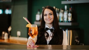 Una persona sostiene un cóctel con pajita, lleva una pajarita y está en un bar con estantes llenos de botellas al fondo.
