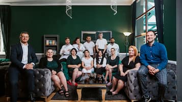 A group of people seated on sofas and chairs in a room with a green wall, a large window, and a chandelier.